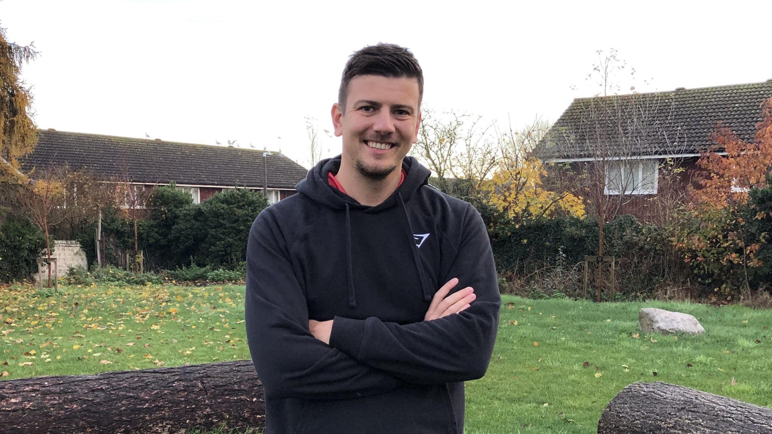 A wider view of the man with short dark hair smiles at the camera. He has a small amount of facial hair and is wearing a black hoodie with a red top underneath. He is standing outside and houses can be seen in the distance behind him.