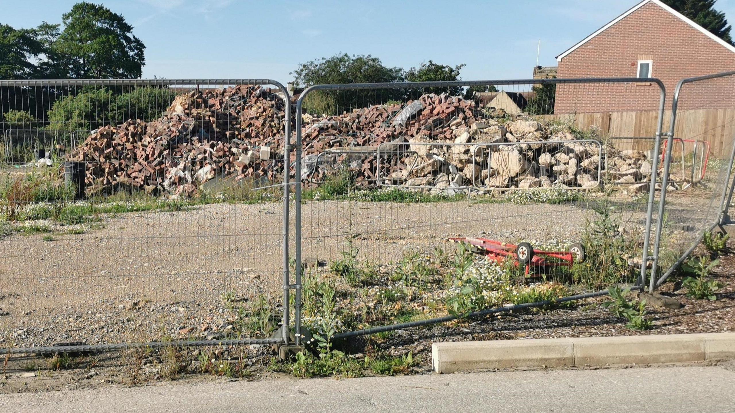 A metal fence in front of a large pile of building rubble. Weeds are growing on the ground. To the right of the rubble is a house.