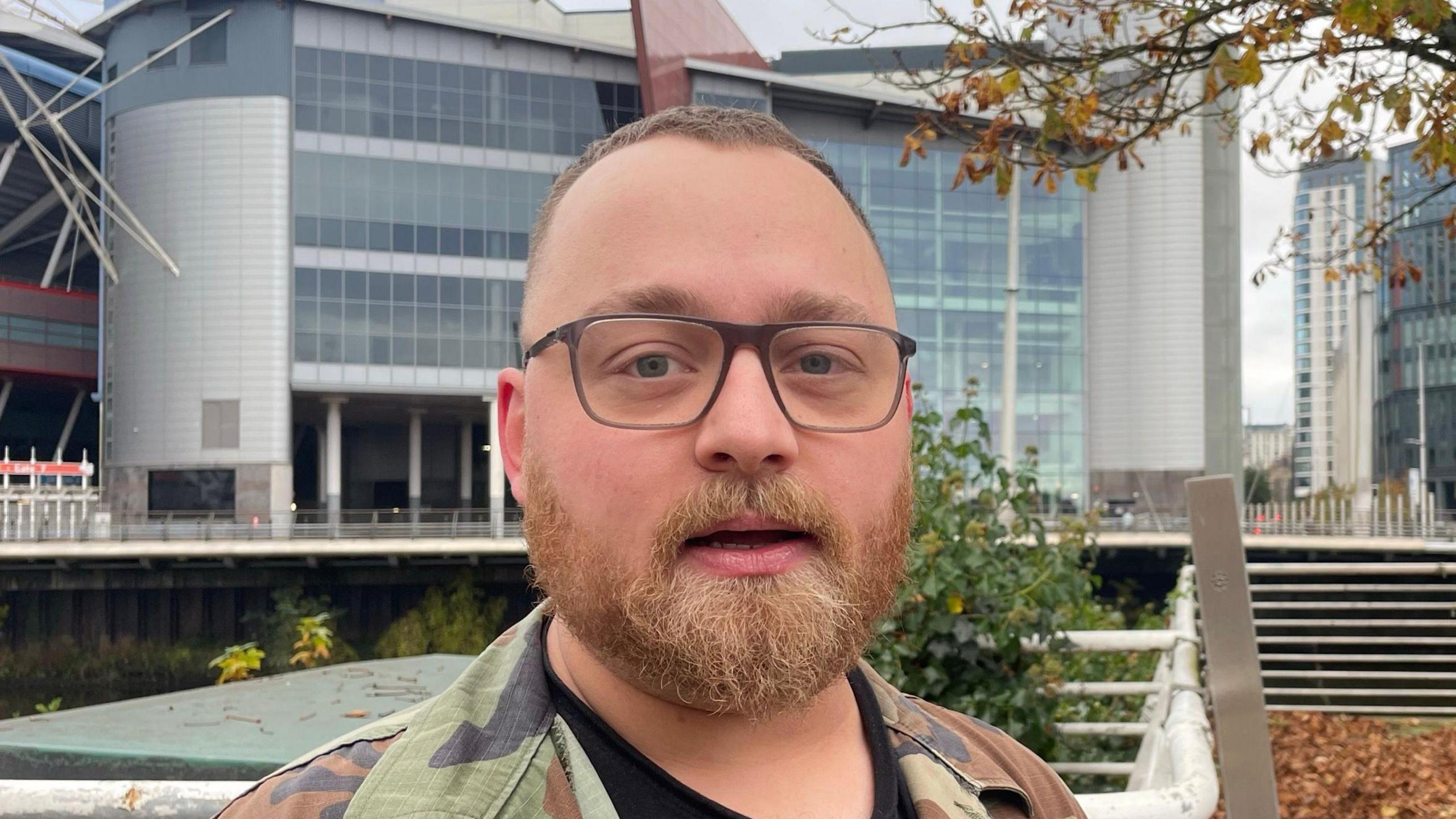 Danny Muir looking at the camera, he has short hair, is wearing black glasses and a camoflage green jacket. He is standing in front of the bridge at Wood Street at the Fitzhamon Embankment. 