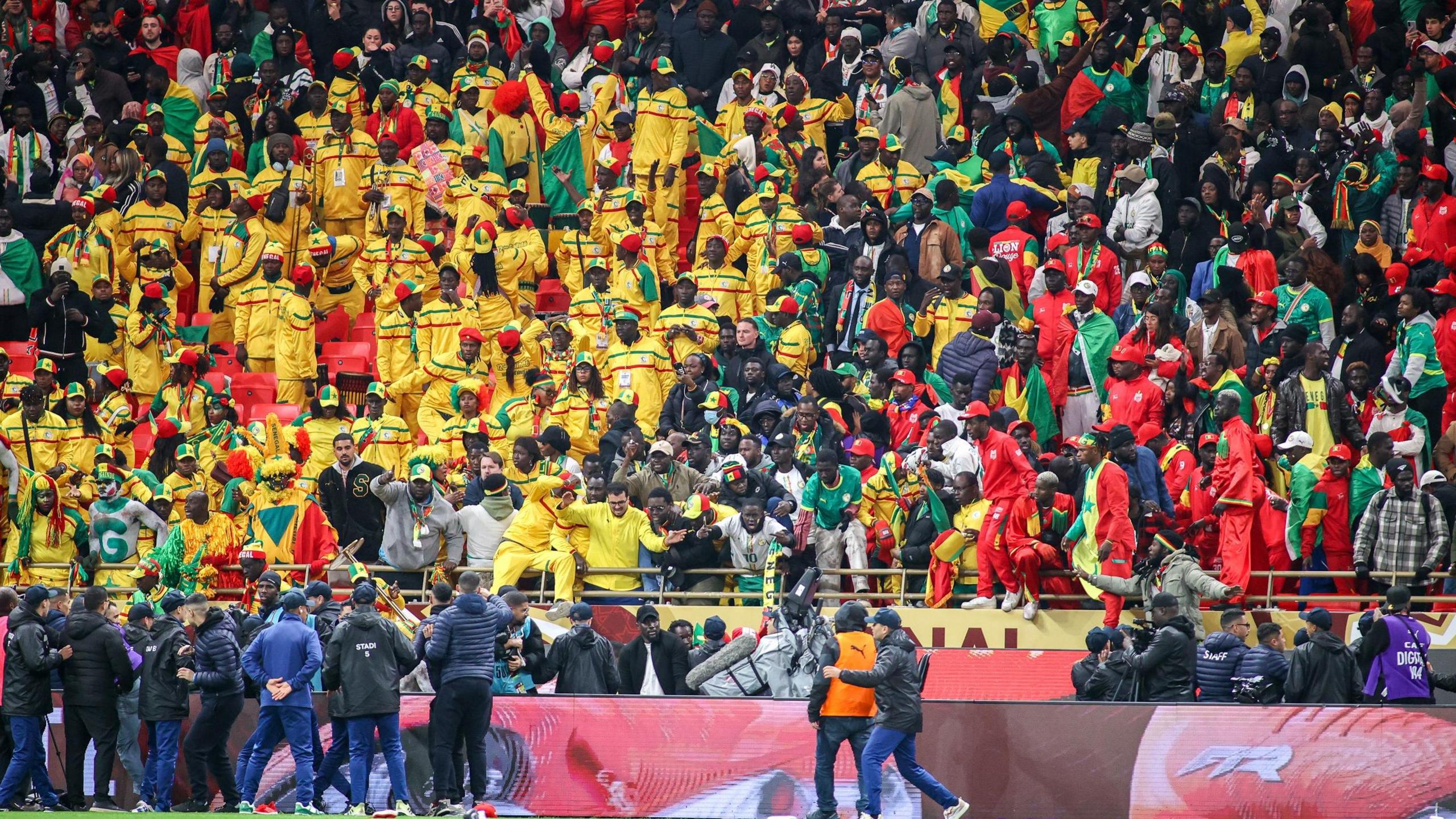 Senegal fans, all dressed in yellow, green and red, are seen in a stand during the 2025 Africa Cup of Nations final. Many are shouting and gesturing down towards security staff who are standing on the edge of the playing surface looking up at them