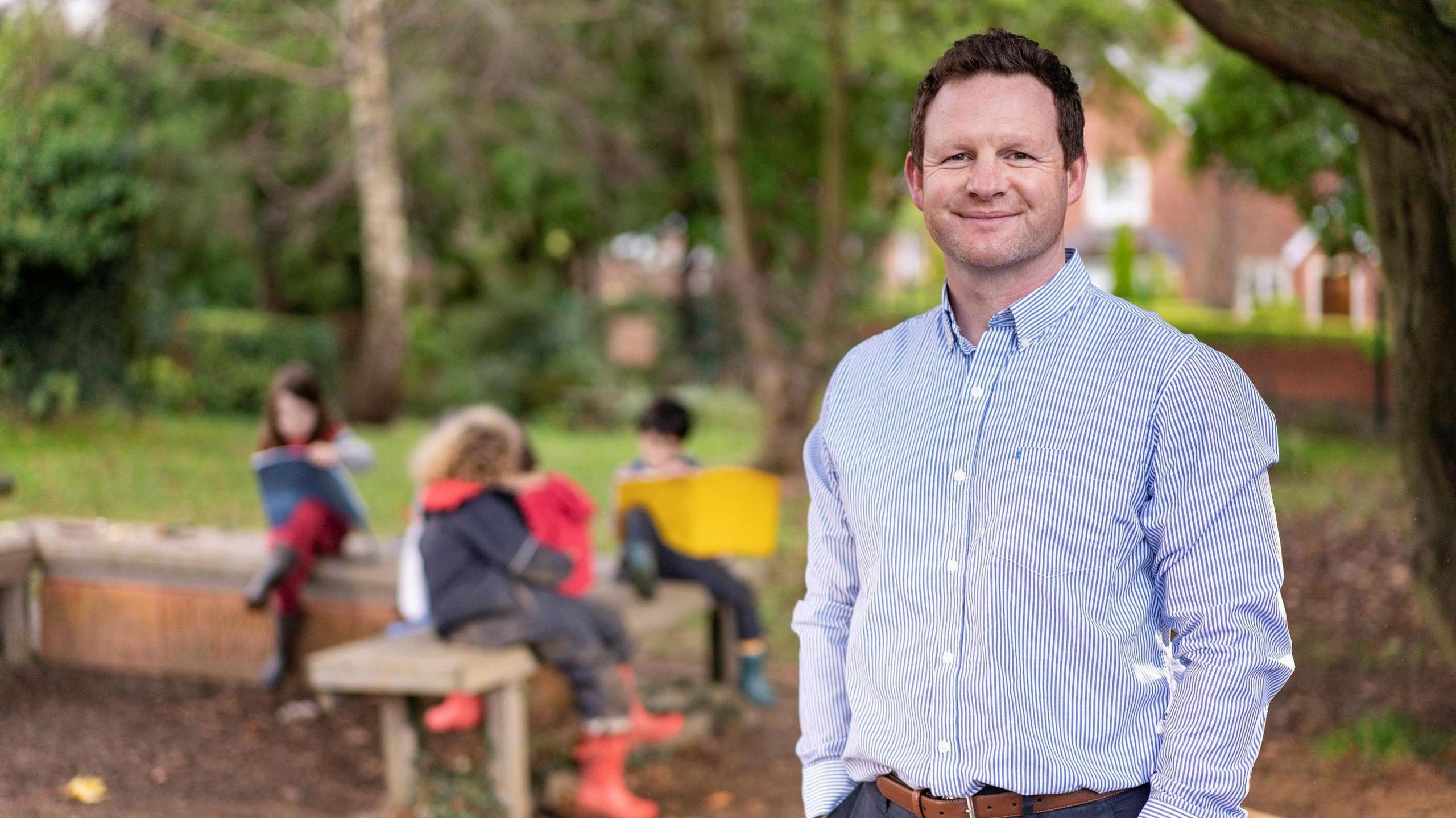 a man in a shirt smiling with his hands in his pockets. Behind him, out of focus, some children in waterproof clothes and boots are sitting on a bench and writing or drawing in big exercise books. The photograph is taken outside and some trees are visible in the background