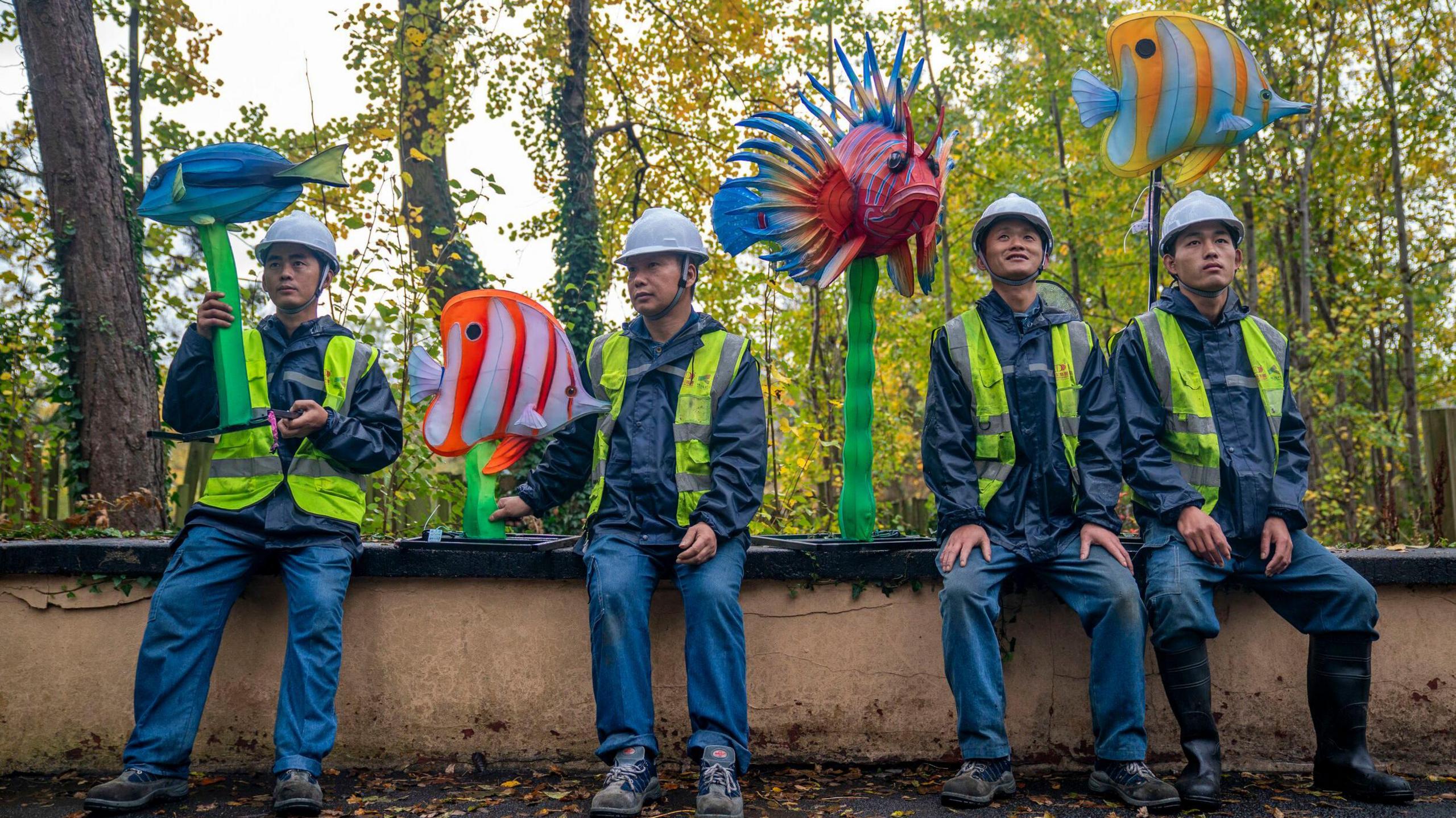 Four men in hard hats and fluorescent jackets sit on a wall carrying lanterns shaped like colourful fish. Trees are in the background