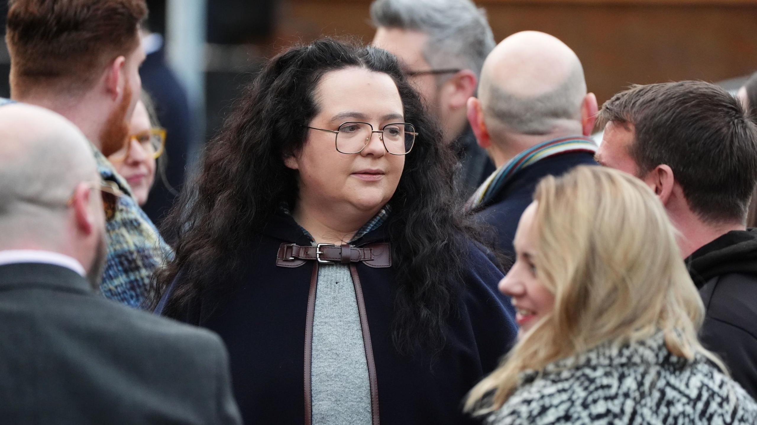 Storrie wearing glasses and a black cape with long curly hair in teh middle of a crowd