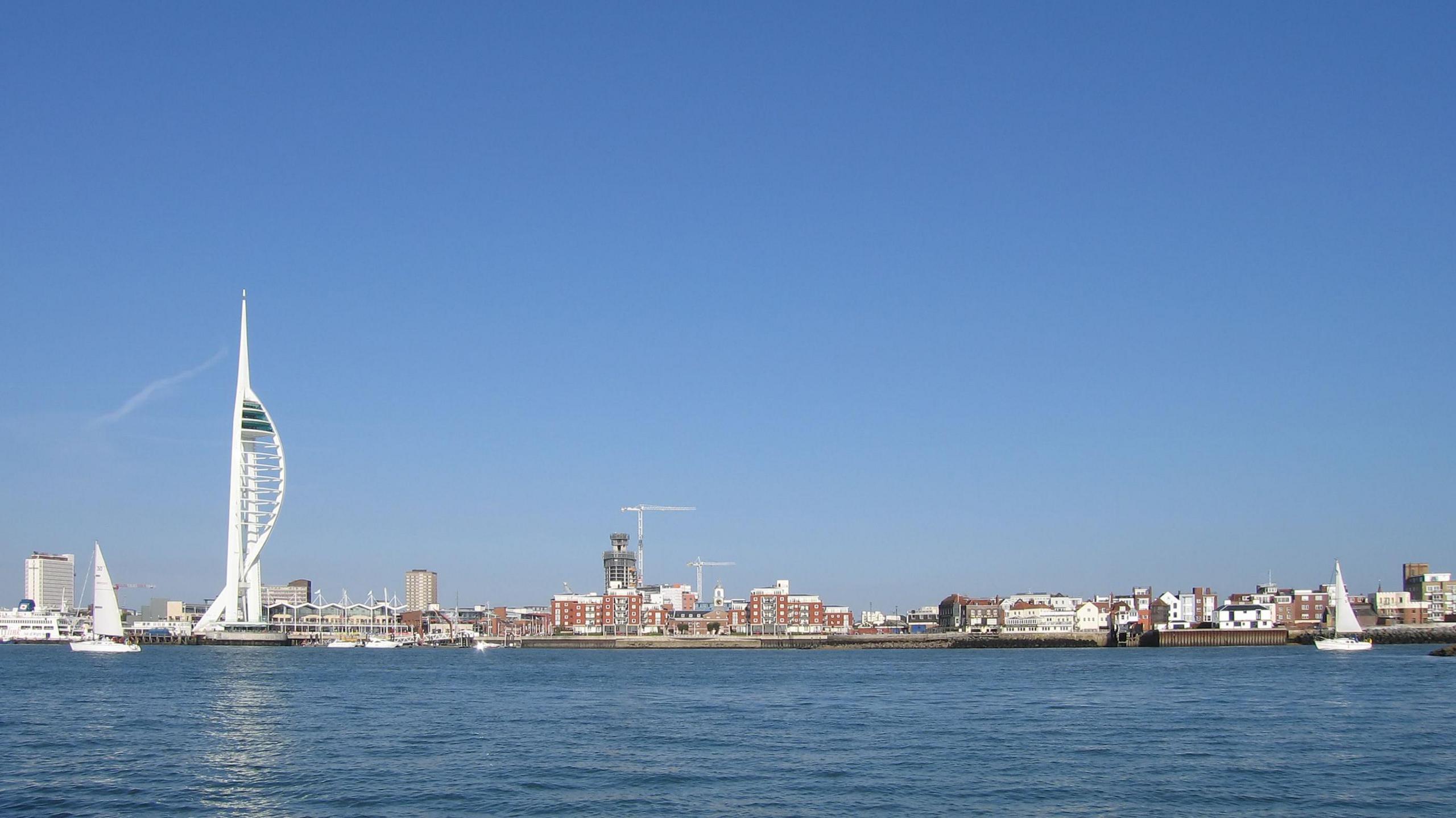 The Portsmouth skyline, taken from out to sea. There's the tall white Spinnaker Tower on the left, some shorter brick buildings in the middle with cranes behind them, and some white buildings on the right.