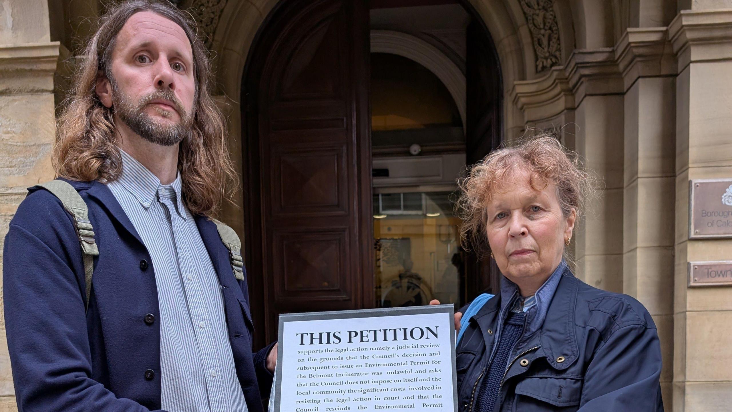 A man and a woman hold up a piece of paper between them which reads, 'THIS PETITION' in capital letters. The writing underneath is illegible. They are stood at the entrance of a town hall.