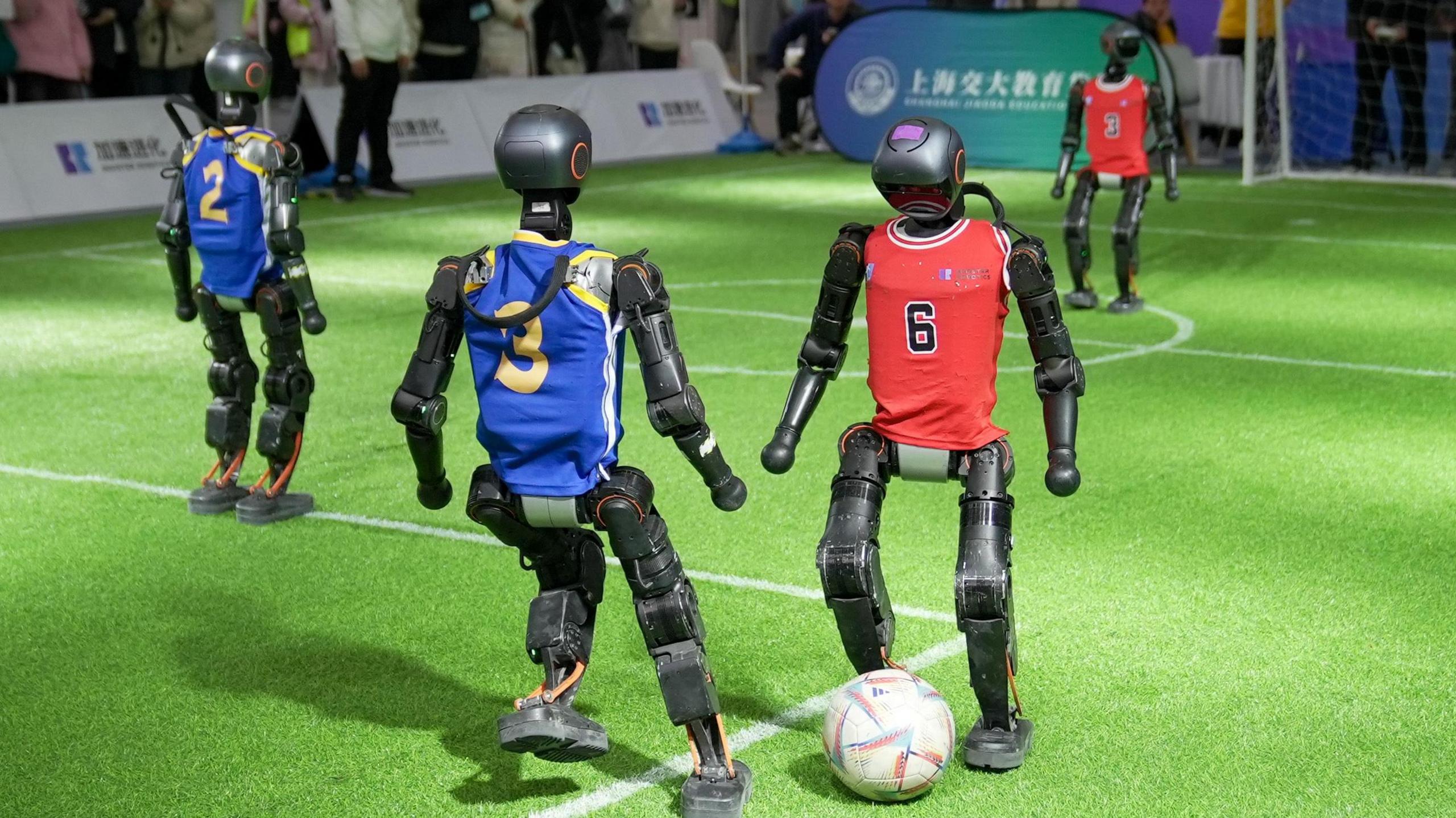 Black robots in blue and red jerseys play football on a green carpet of artificial grass.