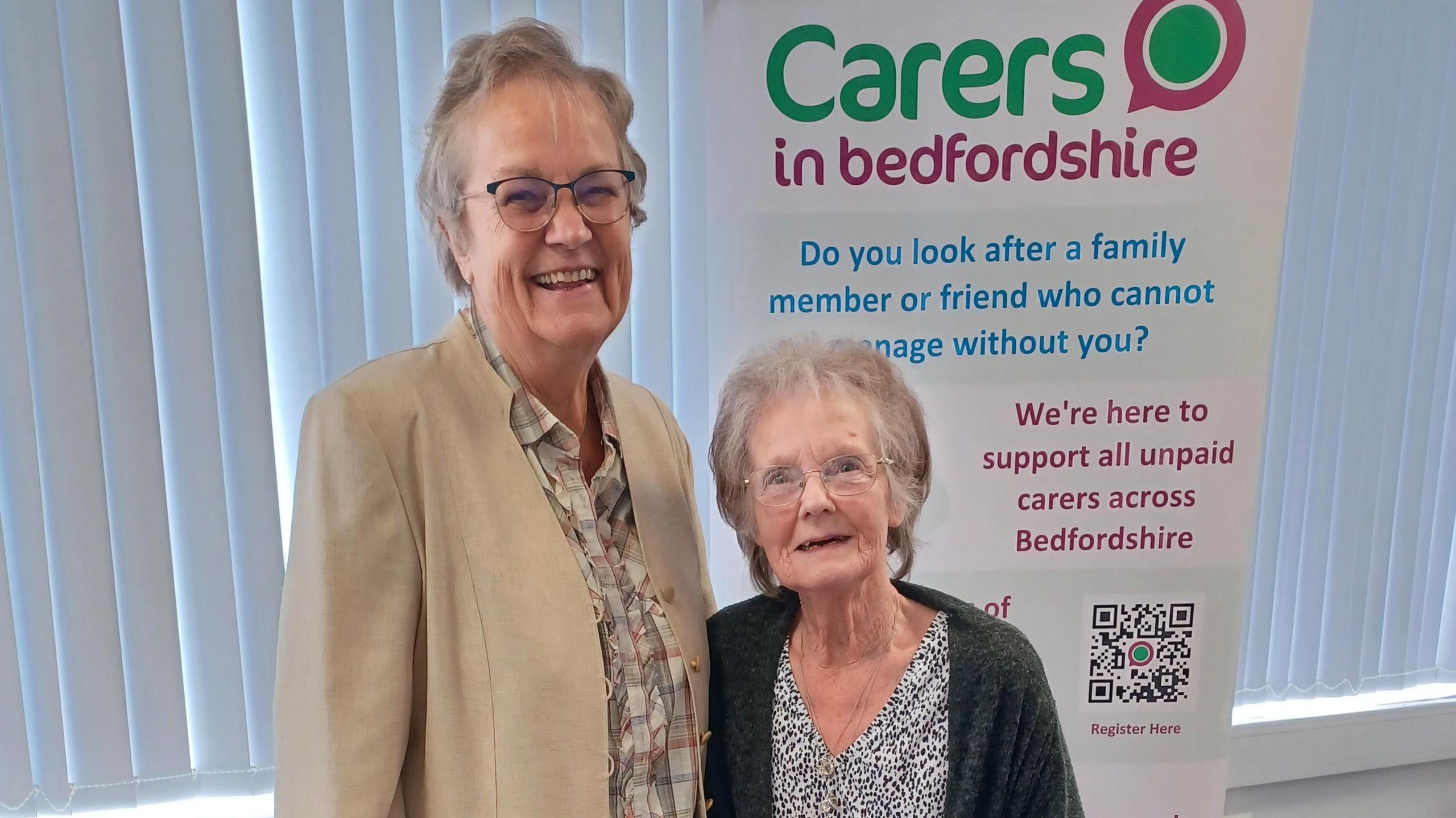 Anne Grant and Yvonne Clark, standing in front of a Carers in Bedfordshire sign, which reads: "Do you look after a family member of friend who cannot manage without you?" Mrs Grant is wearing a checked top and a beige jacket, Mrs Clark is wearing a leopard print top and dark cardigan. They are both looking straight at the camera and smiling.