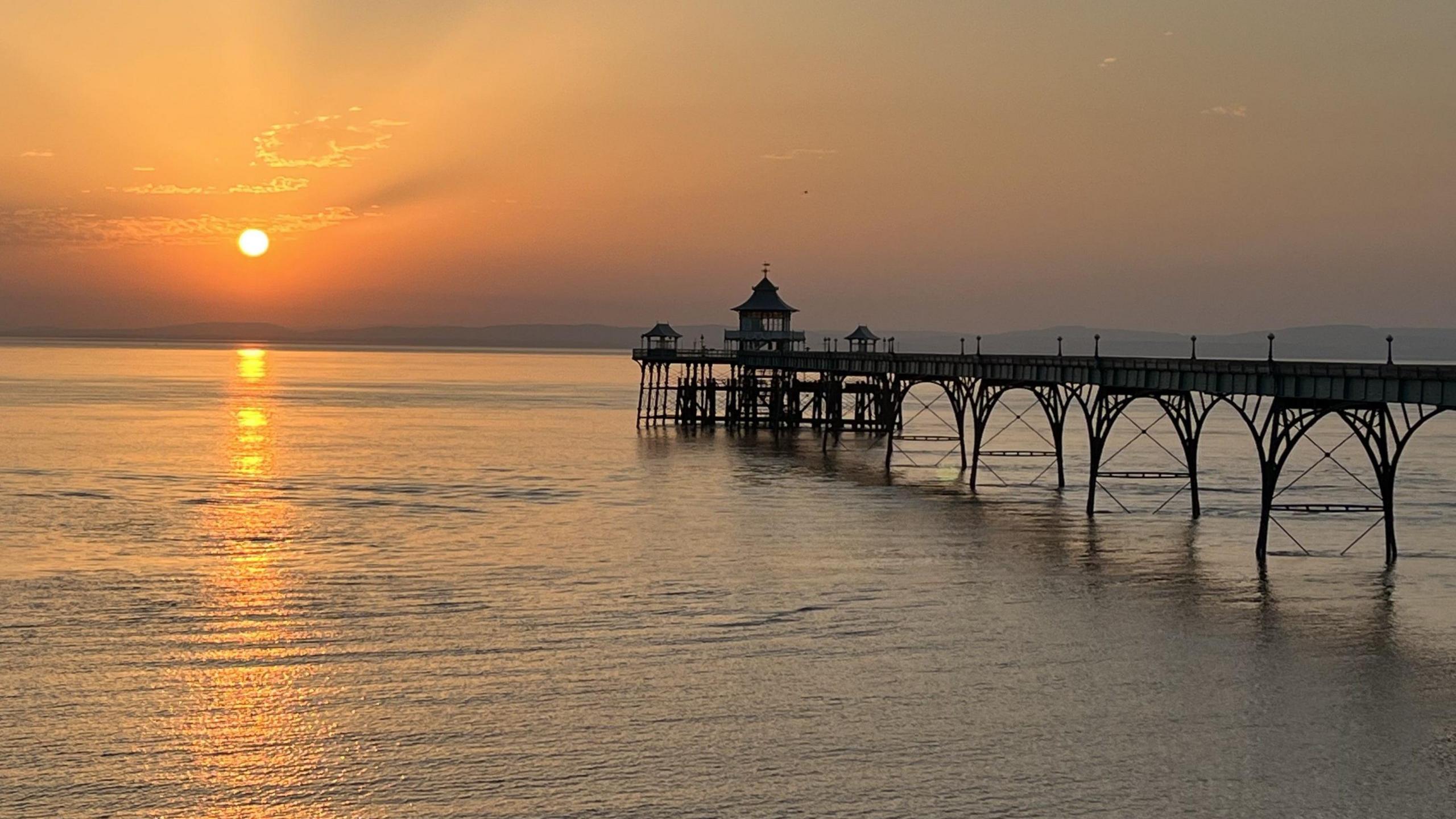 Clevedon Pier is seen silhoutted against an orange sky at sunset, in a picture taken from the shore. The setting sun is a white circle on the left-hand-side of the picture