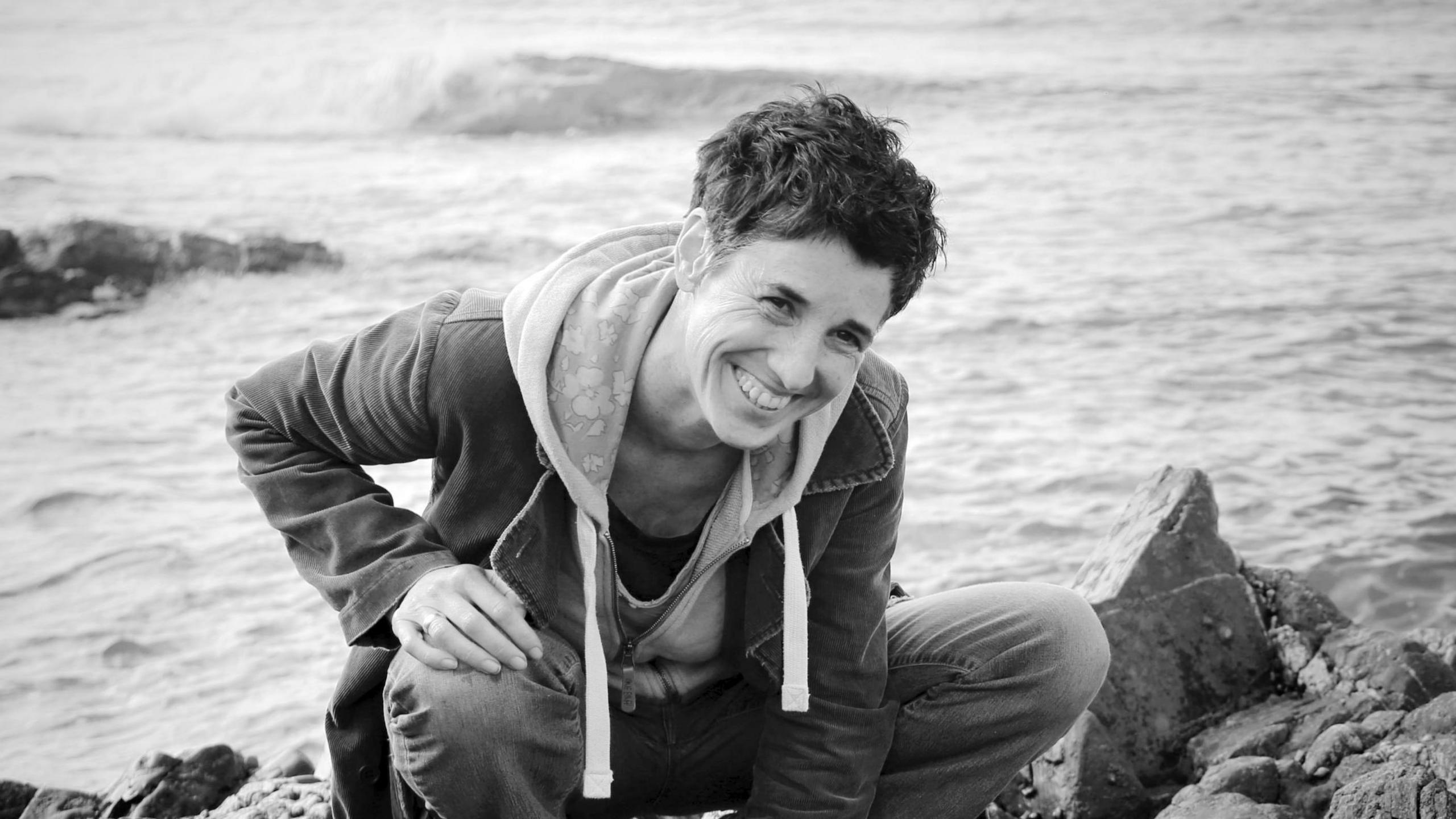 A black and white image of a woman with short dark hair wearing a hoodie and jeans crouching on some coastal rocks. She is smiling and the sea can be seen in the background. 