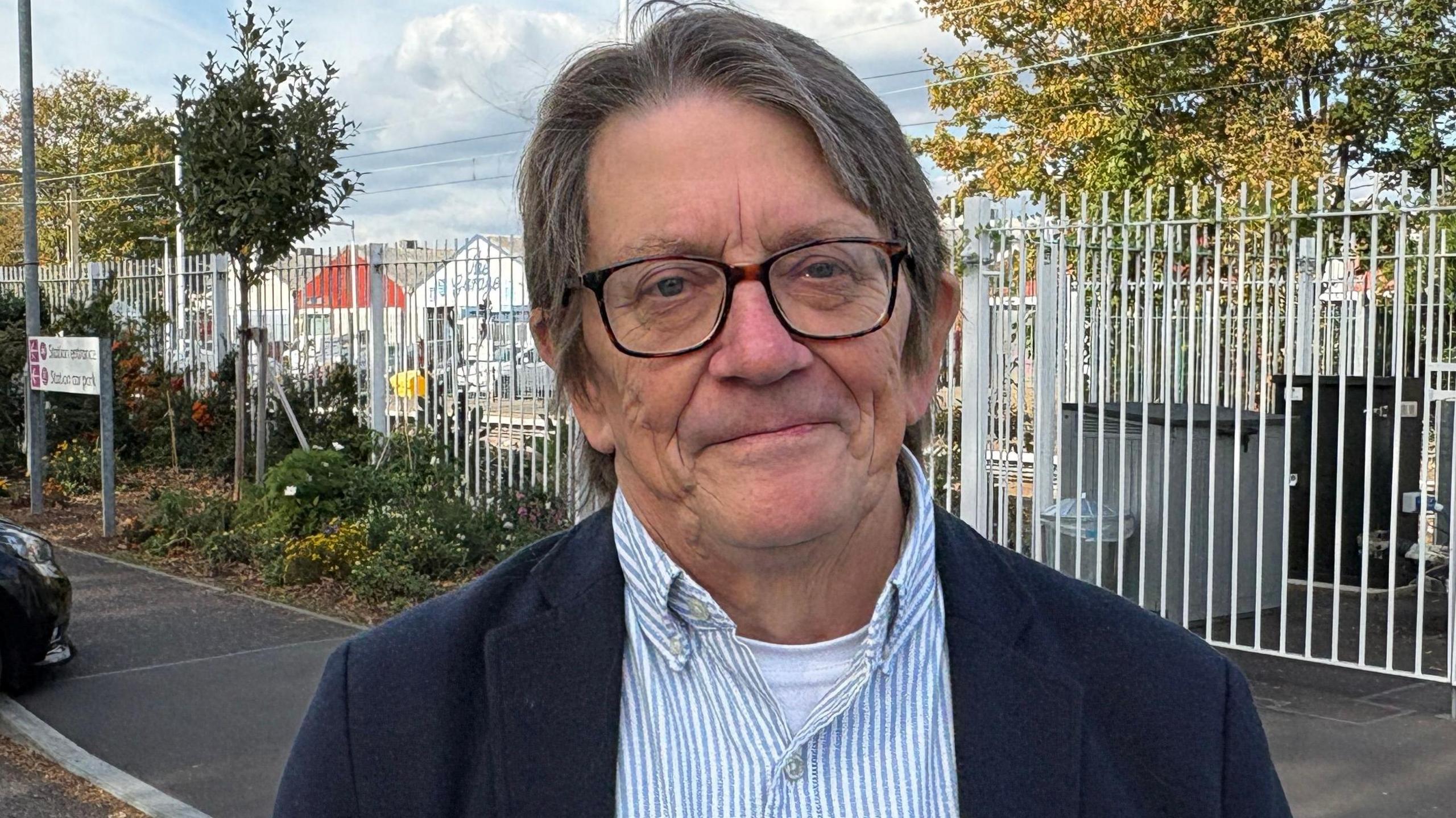 Len Shackleton looking directly down the camera. He is wearing a blue and white striped shirt, with a white t-shirt underneath, and a dark blue jacket on top. He is also wearing rectangular-framed glasses. He has dark hair, showing some grey, which is parted in the middle and falls over his ears. He is standing outside. In the background are tall, white railings, a black tarmac path, and some plants and trees. The front of a car is just visible on the left of frame.