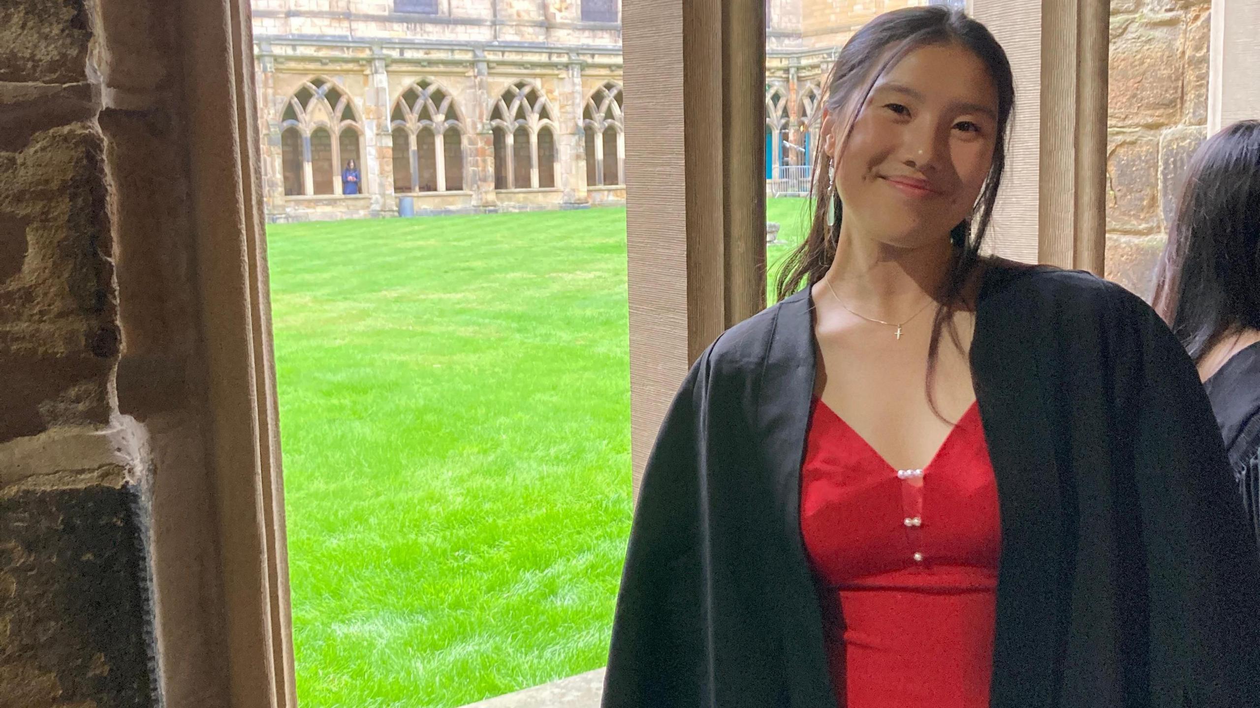 Tian is standing in a red dress in a cathedral courtyard. She is wearing black robes and is smiling at the camera.