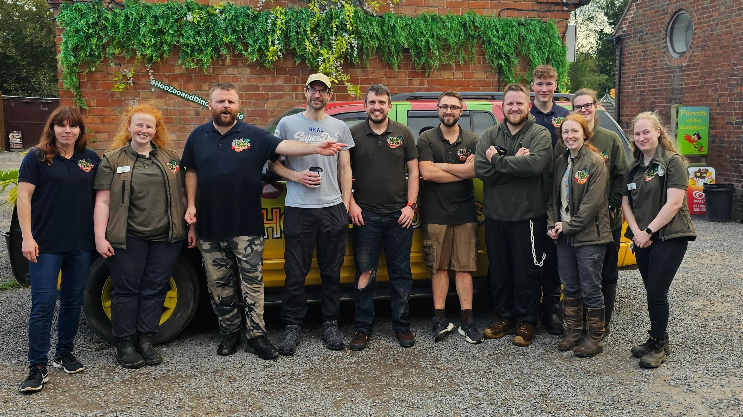 Eleven people standing in front of a yellow car in clothing with Hoo Zoo badges, all smiling. They are outside in a carpark in front of a wall.