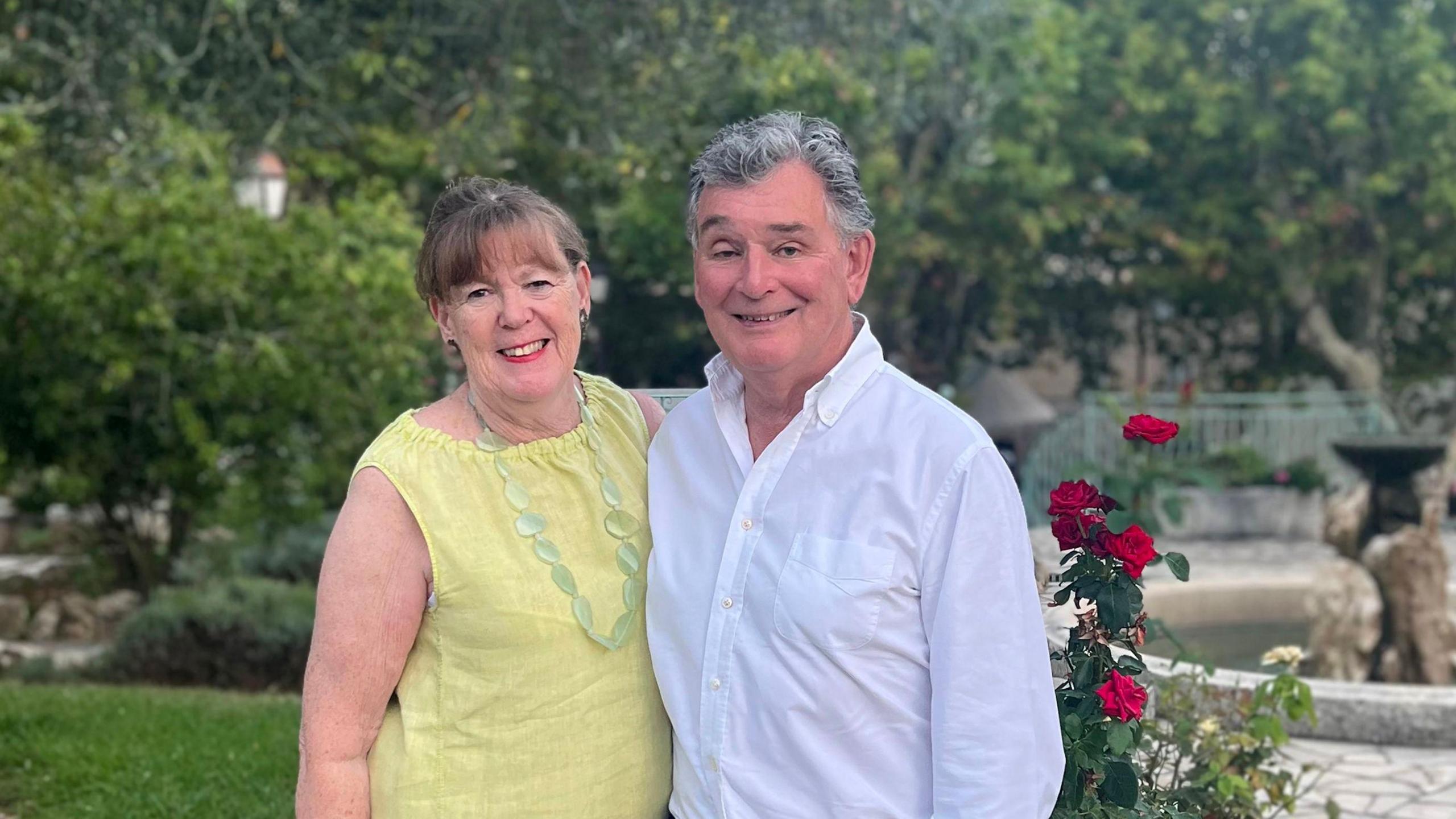 A photo of Judy and Jonathan Bloomer. They are standing together with their arms around each other. They are standing in a garden with bushes and flowers behind them. Judy Bloomer is wearing a yellow top and Jonathan Bloomer is wearing a long-sleeved white shirt.