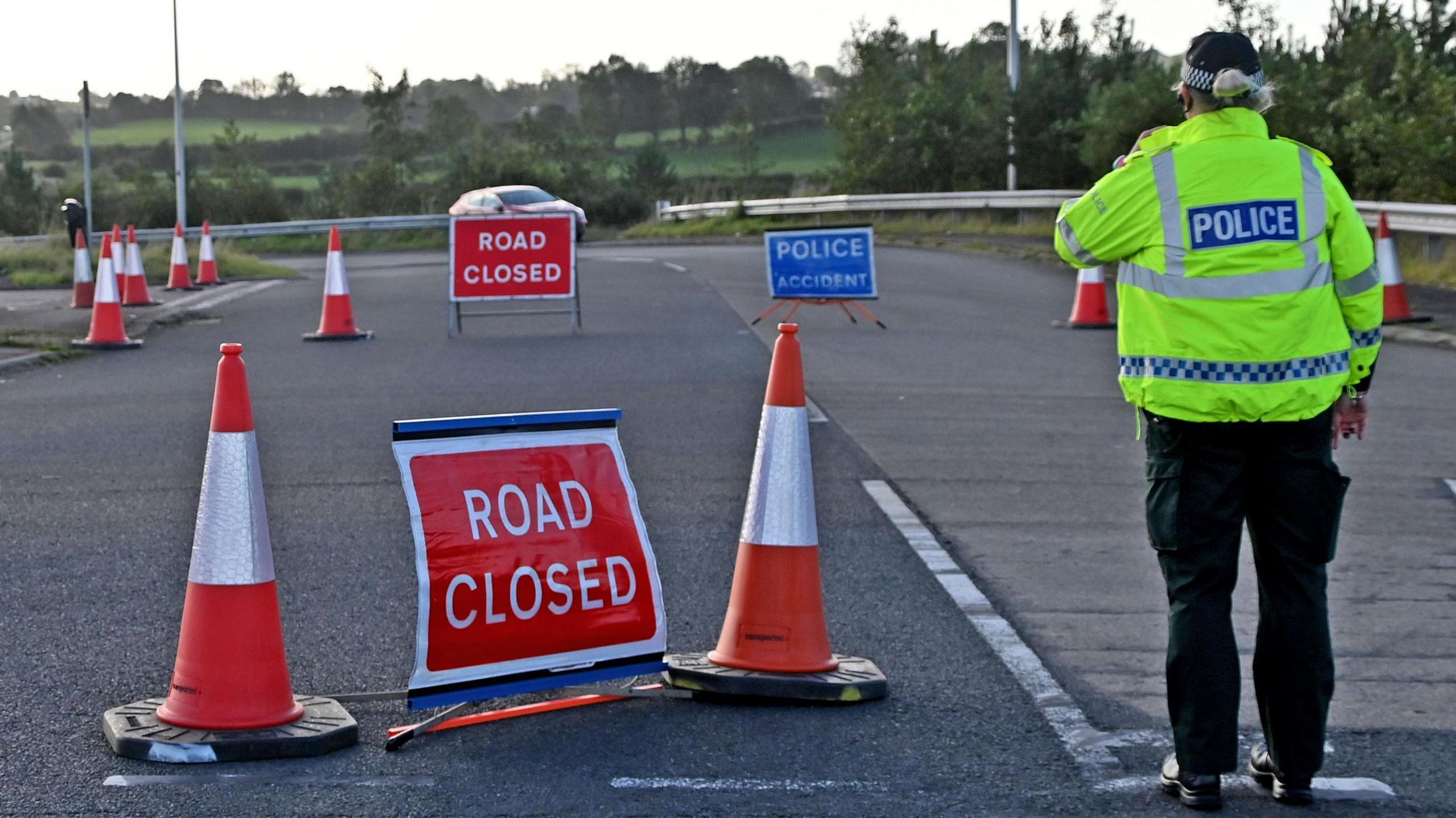 Two red and white traffic cones sit either side of a red and white sign that reads 'Road Closed'. Similar signs are visible in the background. To the right of the sign is a police officer standing with their back to the camera. The officer is wearing dark green trousers, a fluorescent yellow jacket and a dark green cap.