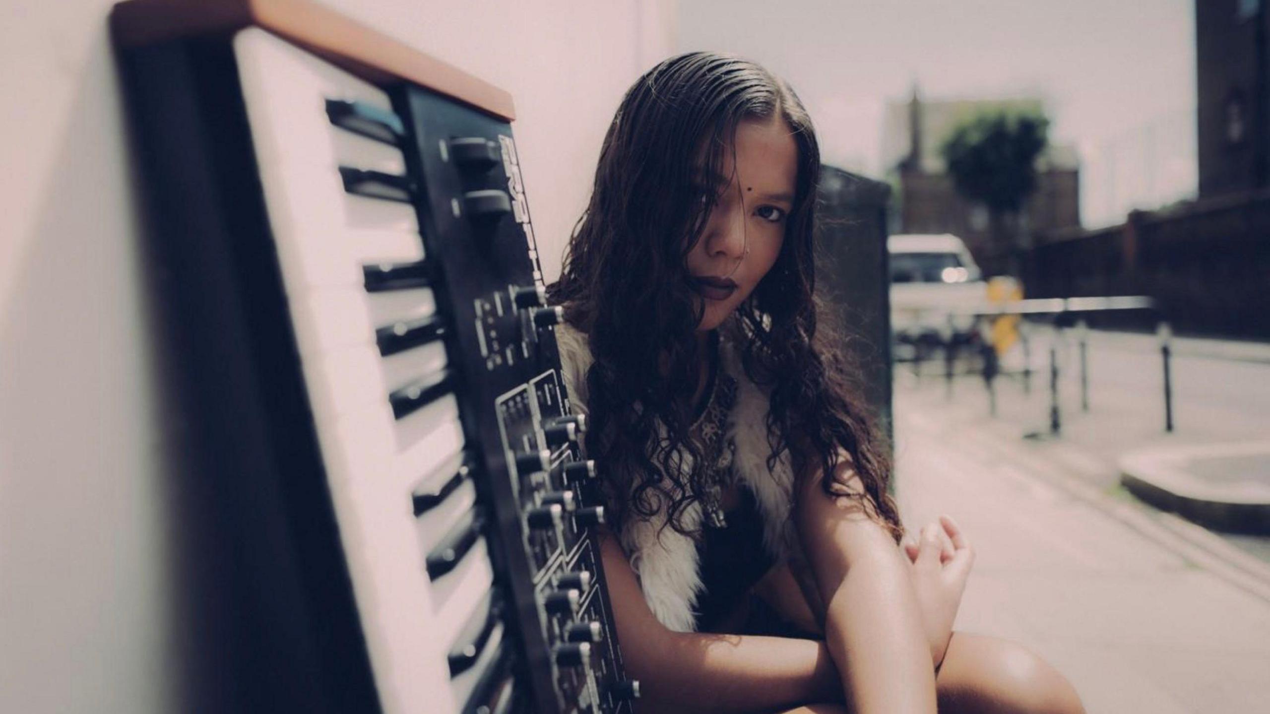 A woman with long, dark hair, sits next to a keyboard which is leaning against a wall on a street