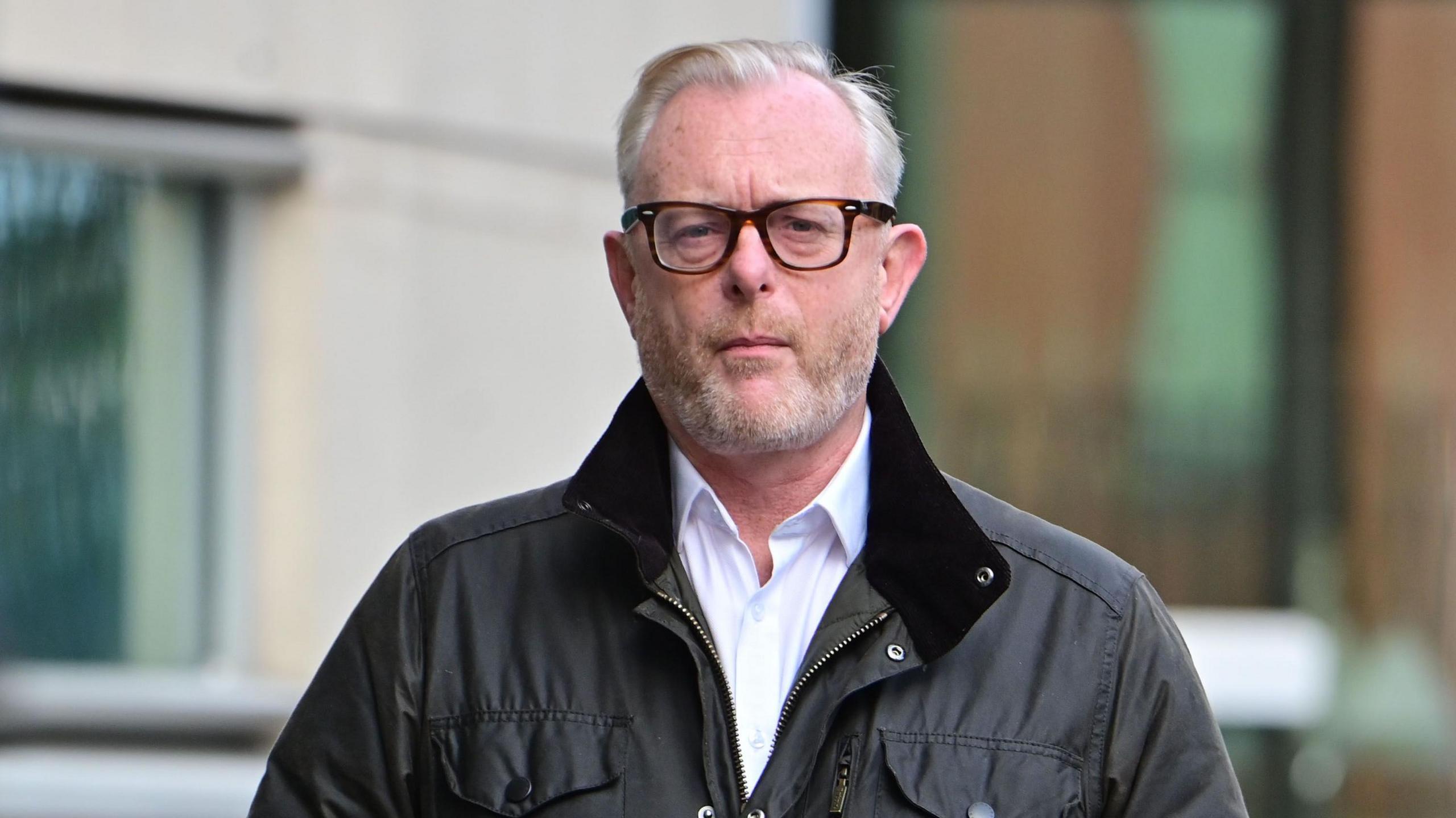 A man with short white hair is pictured walking outside a building. He is wearing dark rimmed glasses, a white shirt, blue jumper, and dark jacket. Behind him is a glass door.