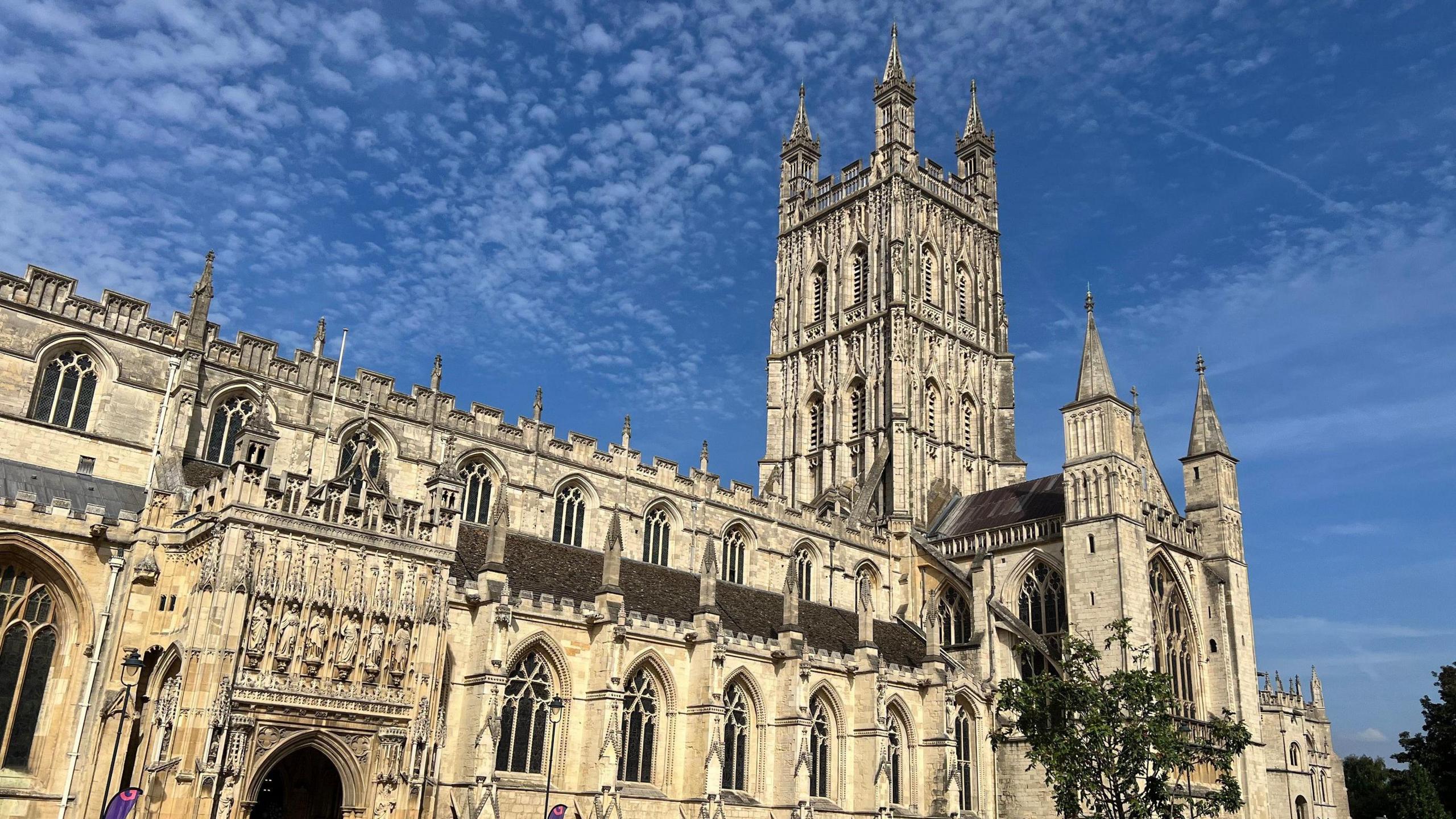 Gloucester Cathedral on a sunny day, with lots of small clouds, almost polka dotted across the blue sky above. A tree can be seen in the foreground.