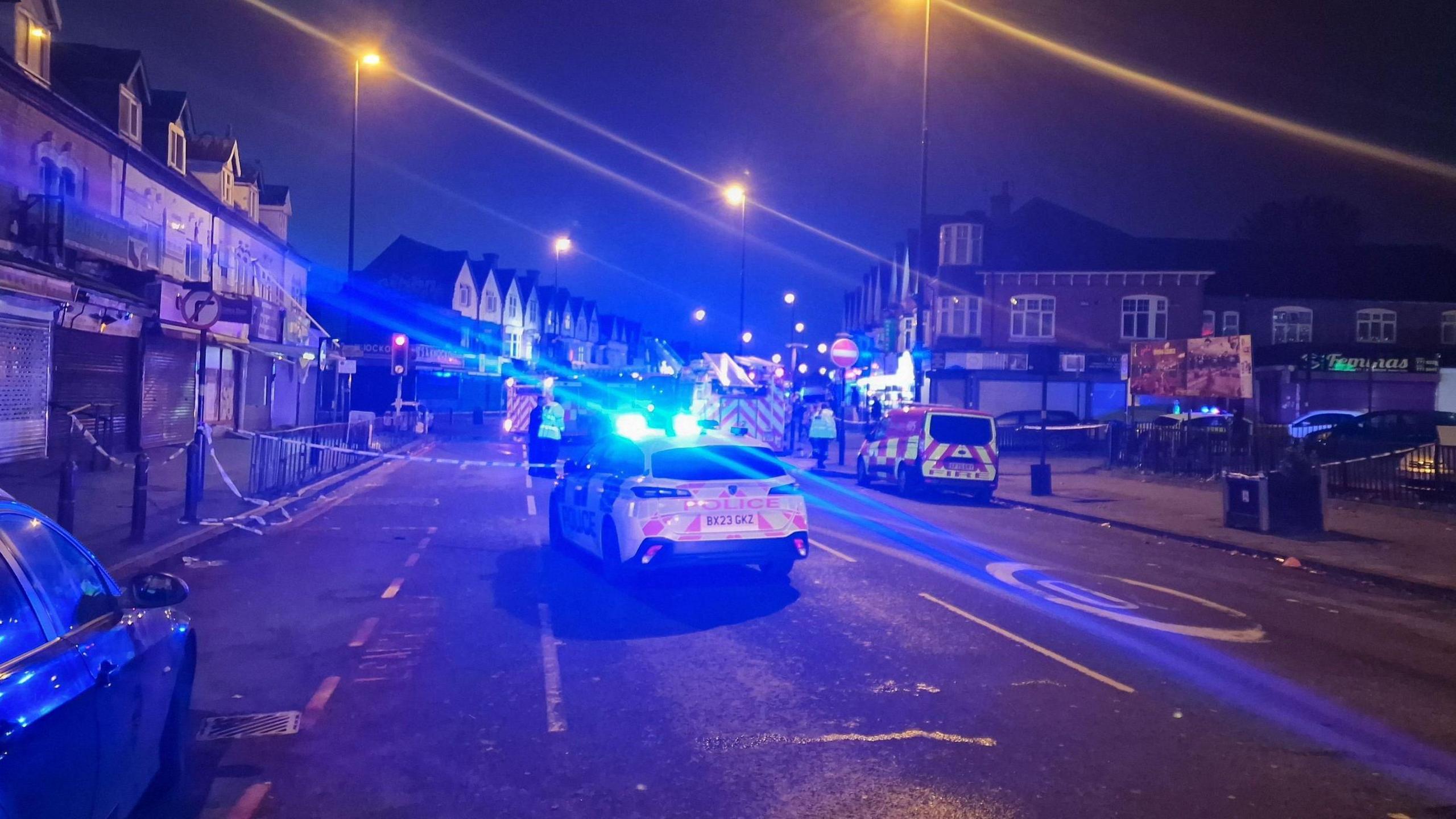 A police car with blue lights is parked in the middle of the road and there is blue and white police tape visible. It is still dark and the blue lights are reflected across the image.