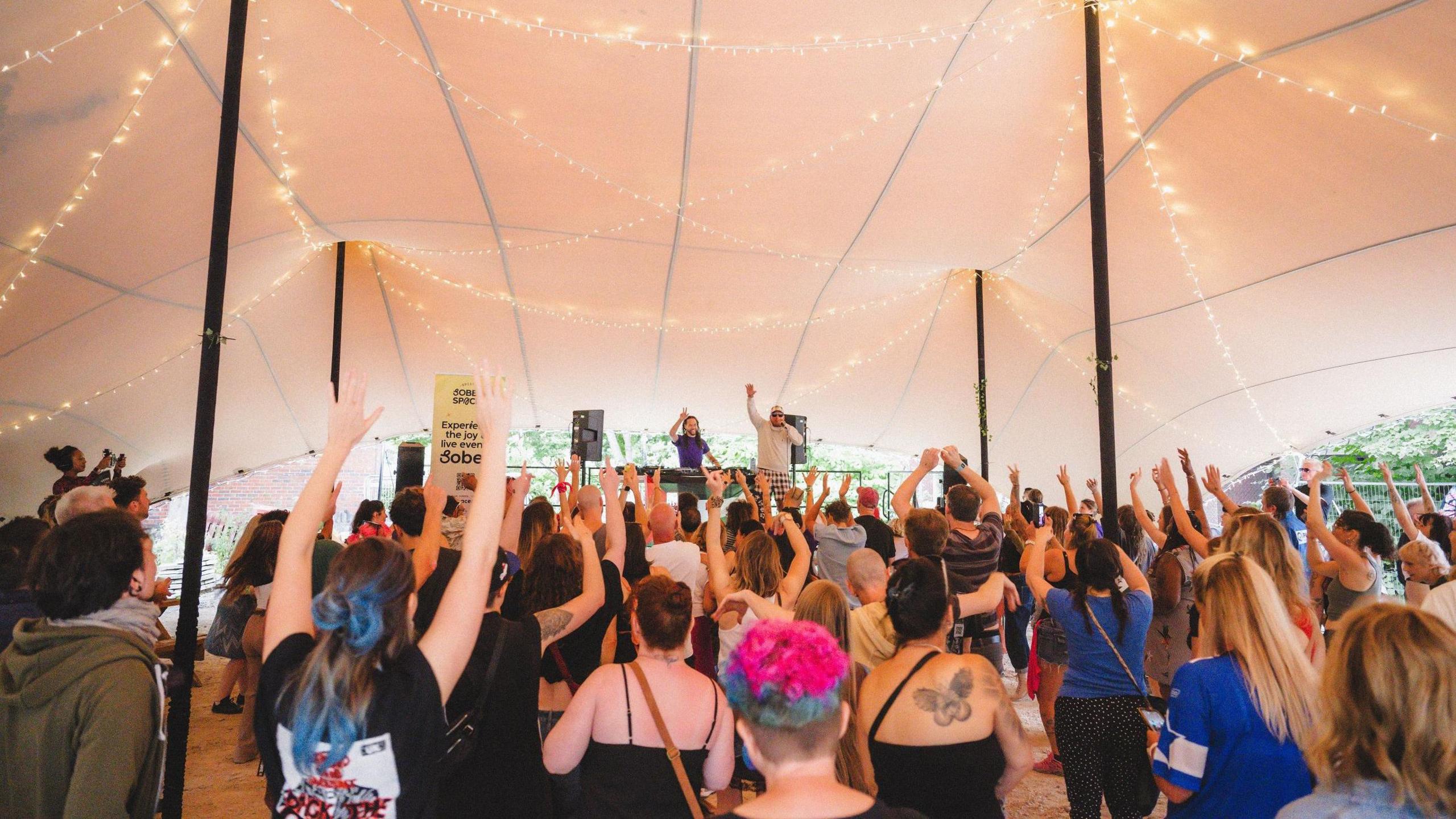 A crowd throwing their hands up in the air as they watch the DJ Roni Size performing on a stage under a lit canopy. Both Roni and his MC have their hands up in the air.