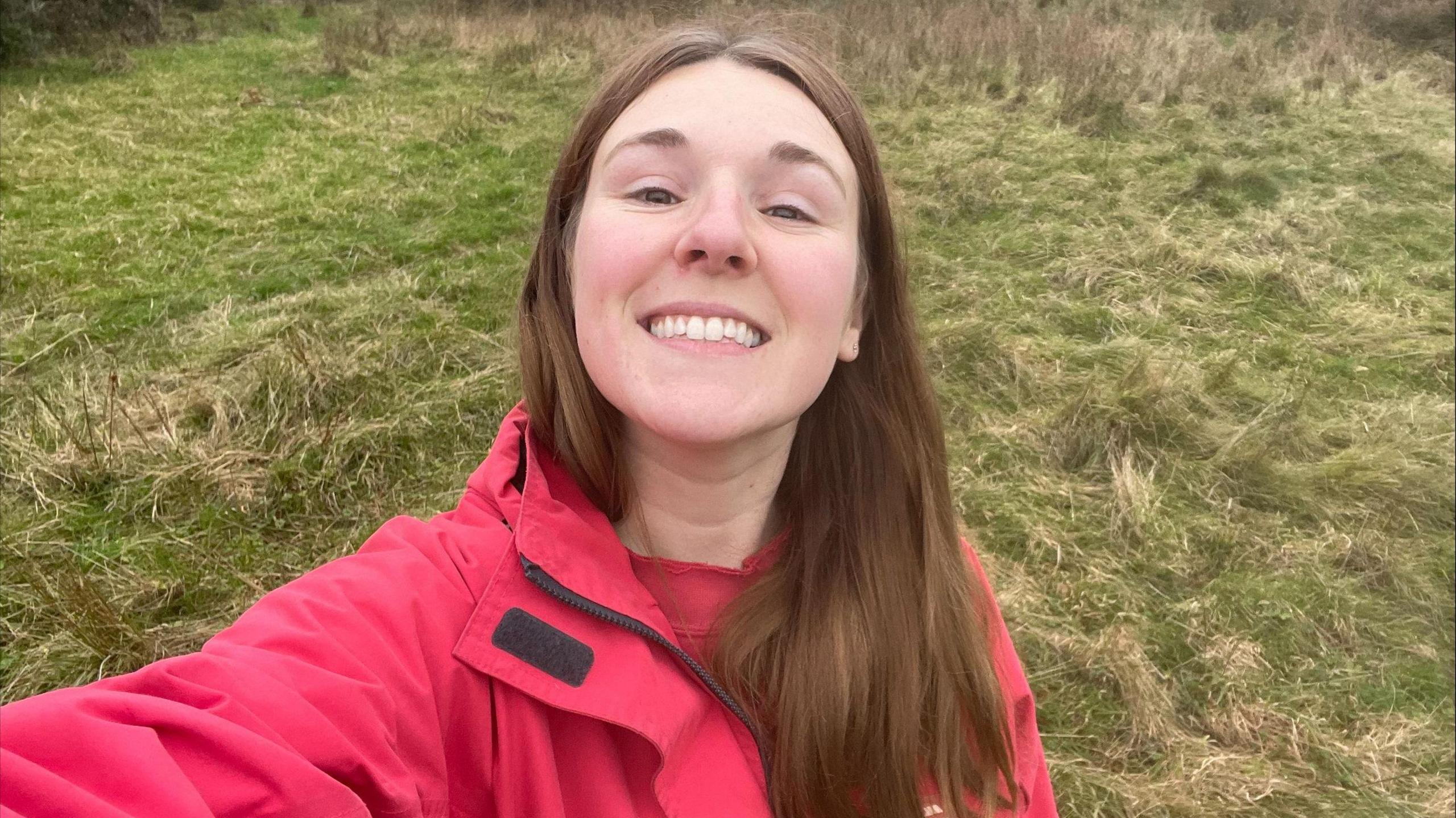 Picture of Becky Davies smiling in a field with her hair worn down behind her ears, wearing a pink raincoat.  