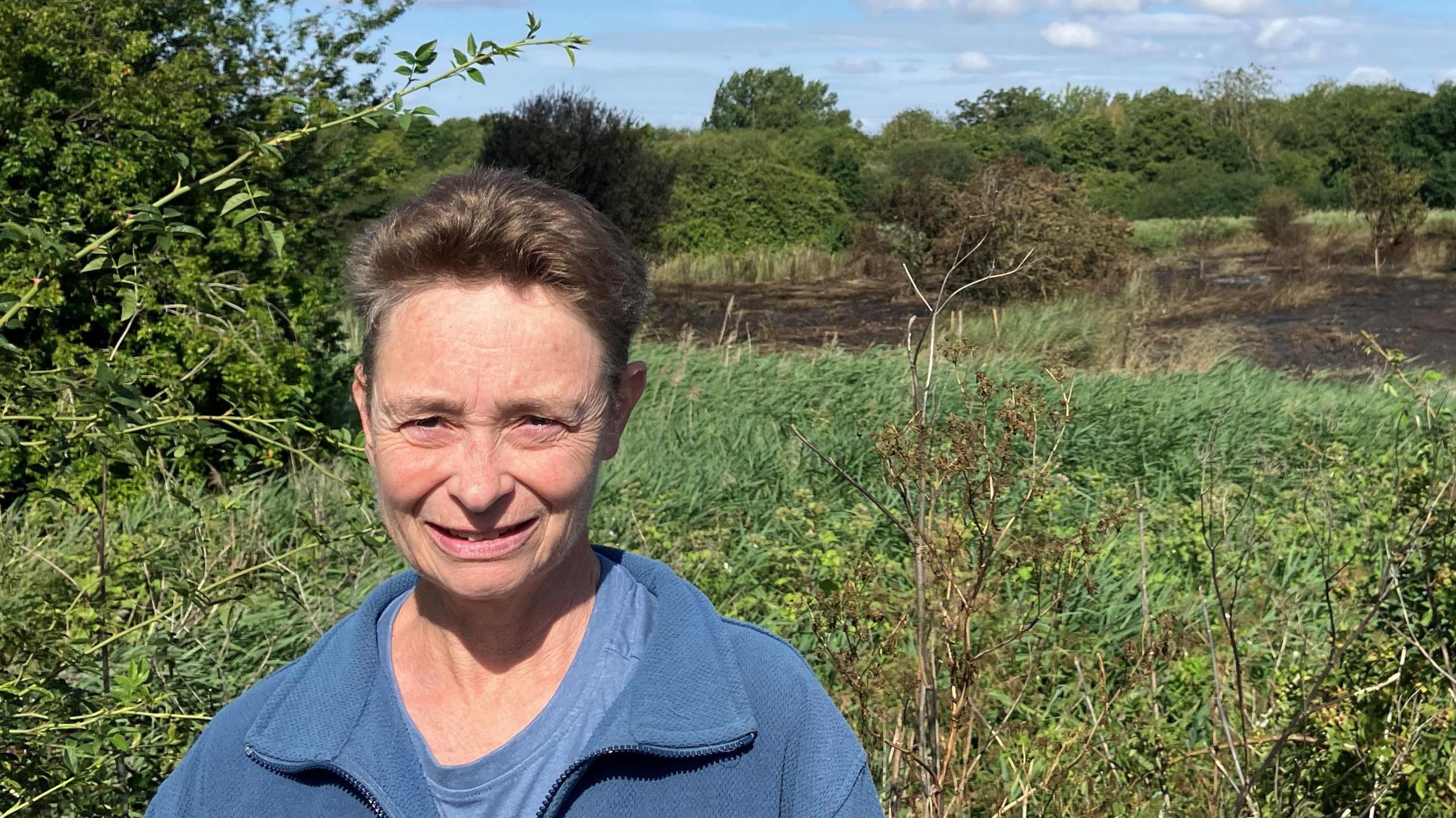A woman in a blue cardigan with lots of greenery in the background, the burned parts of the reserve are visible further back