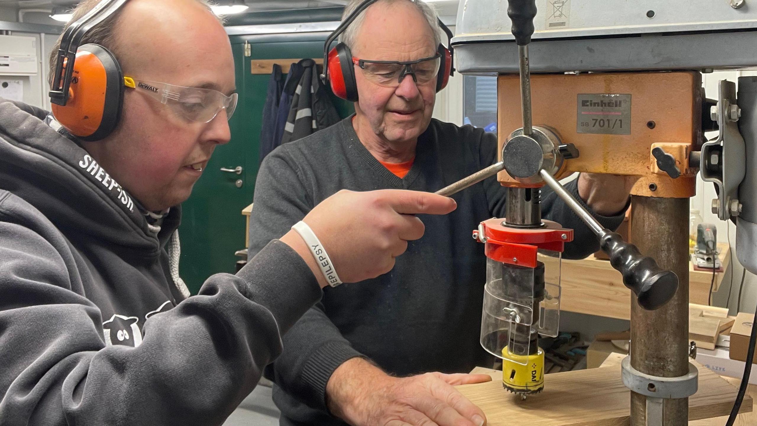 John Huggins, wearing ear protectors and safety goggles, oversees a student, who is also wearing the same safety equipment, pressing down on a machine to drill a hole in wood.