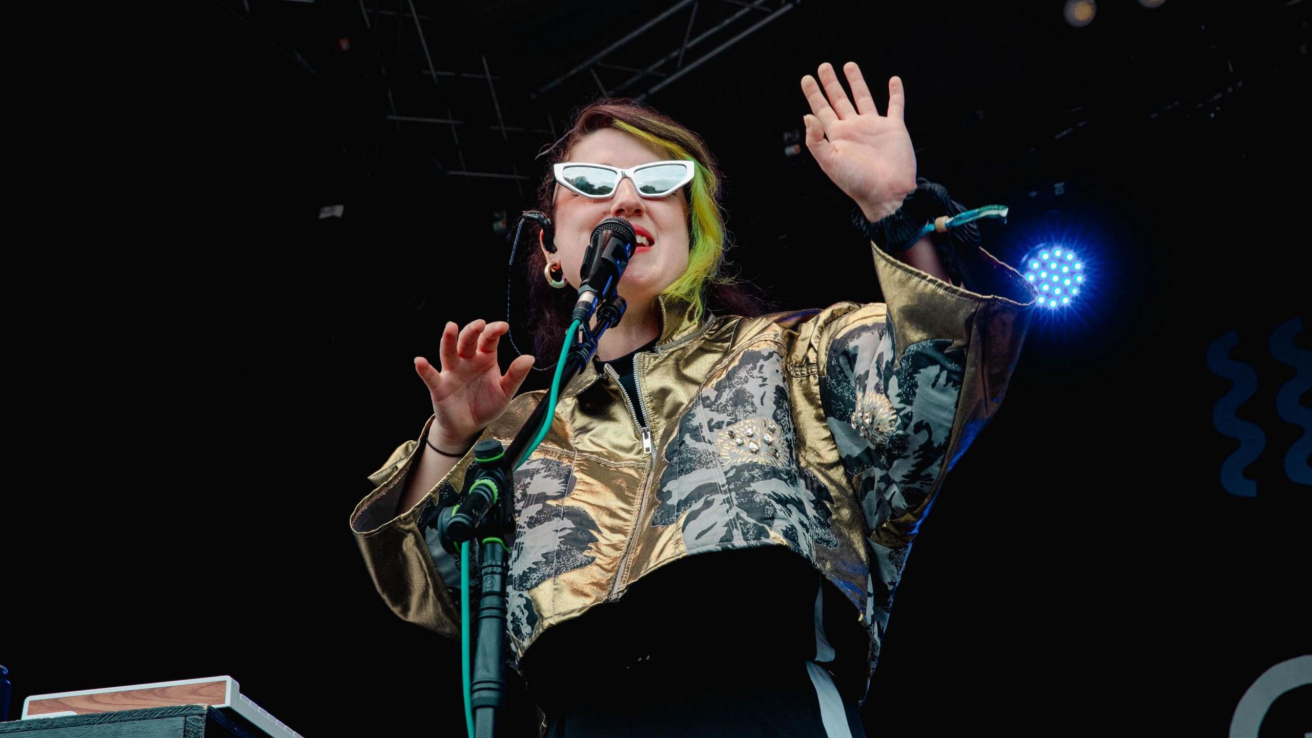 Heledd Watkins, wearing sunglasses and a gold jacket, performs at Green Man