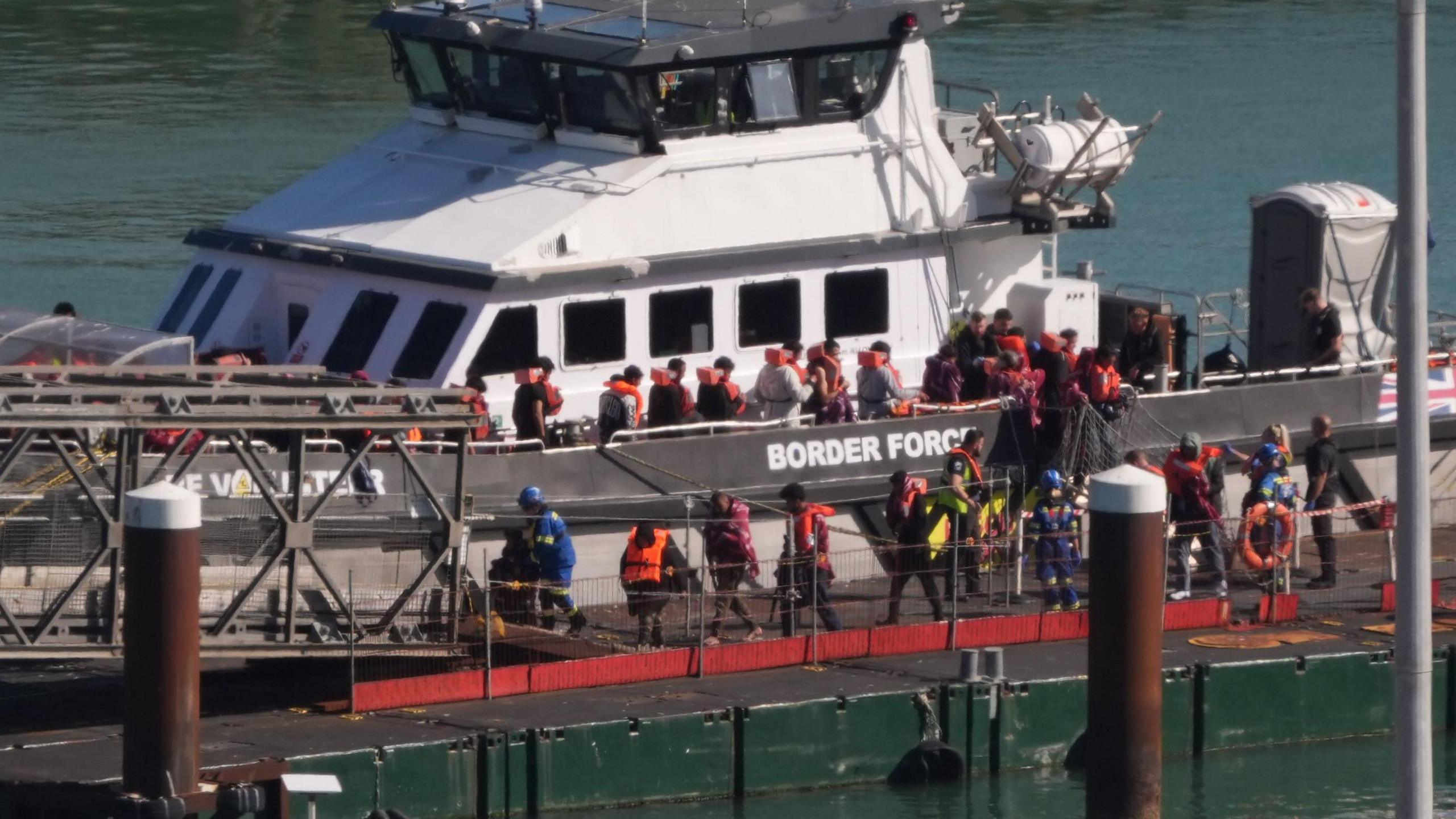 A group of migrants, all wearing red life jackets, disembark from a white and grey border force vessel docked in Dover. They walk down the gang plank and onto the pier in single file.
