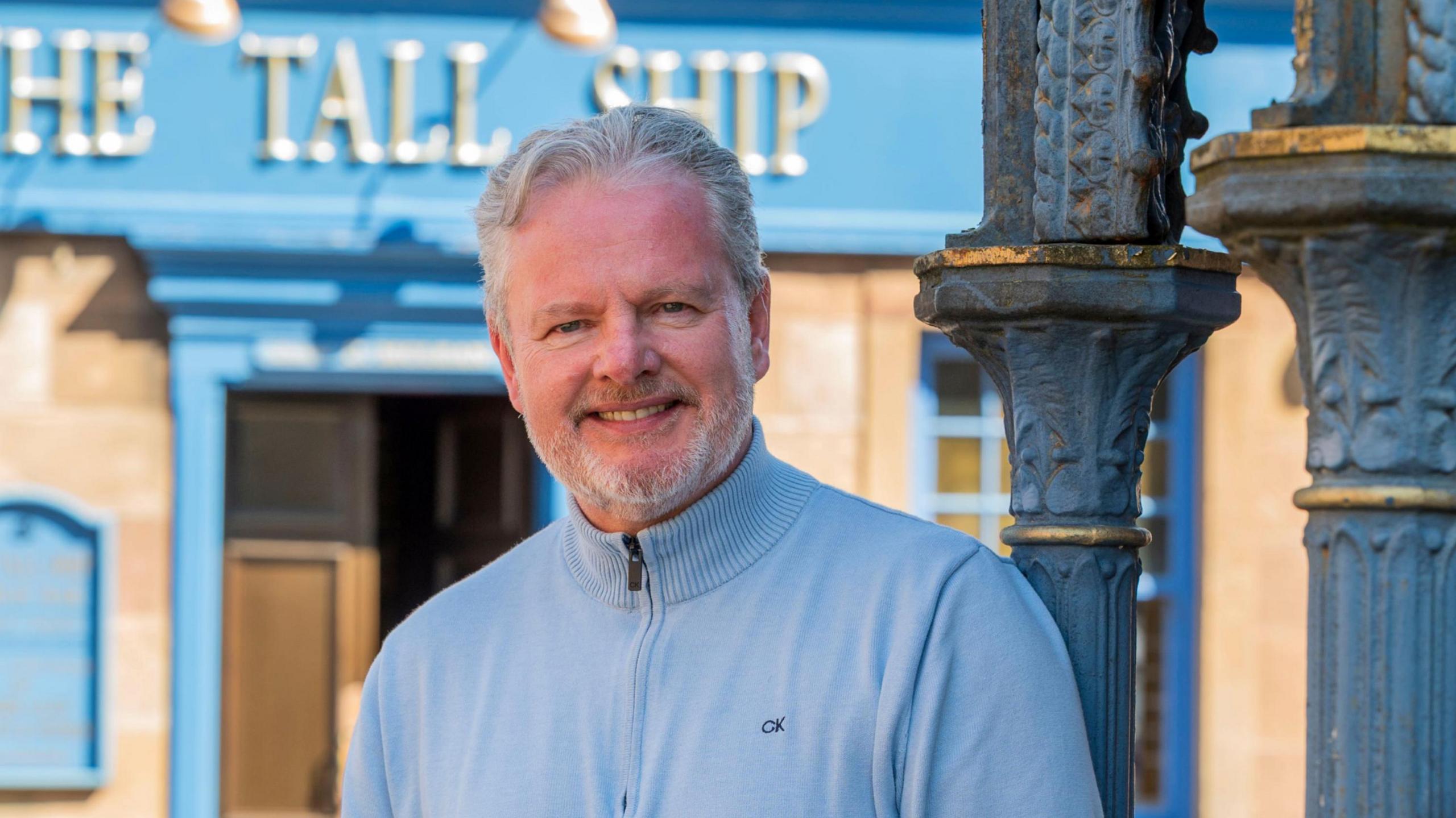 A person stands outdoors beside a decorative cast‑iron column, wearing a light blue sweater and jeans.
Behind them is a stone building with bright blue trim and a sign reading “The Tall Ship” above the doorway.