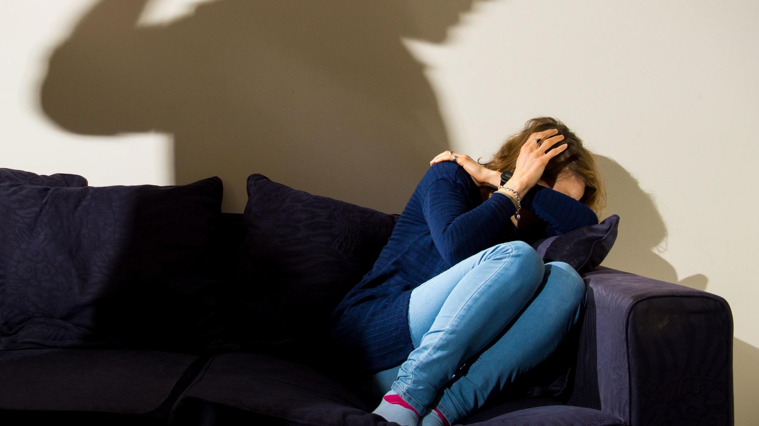 A woman wearing a navy jumper and light blue denim skinny jeans cowers in fear on a navy sofa as the shadow of a man with his fist raised looms over her.