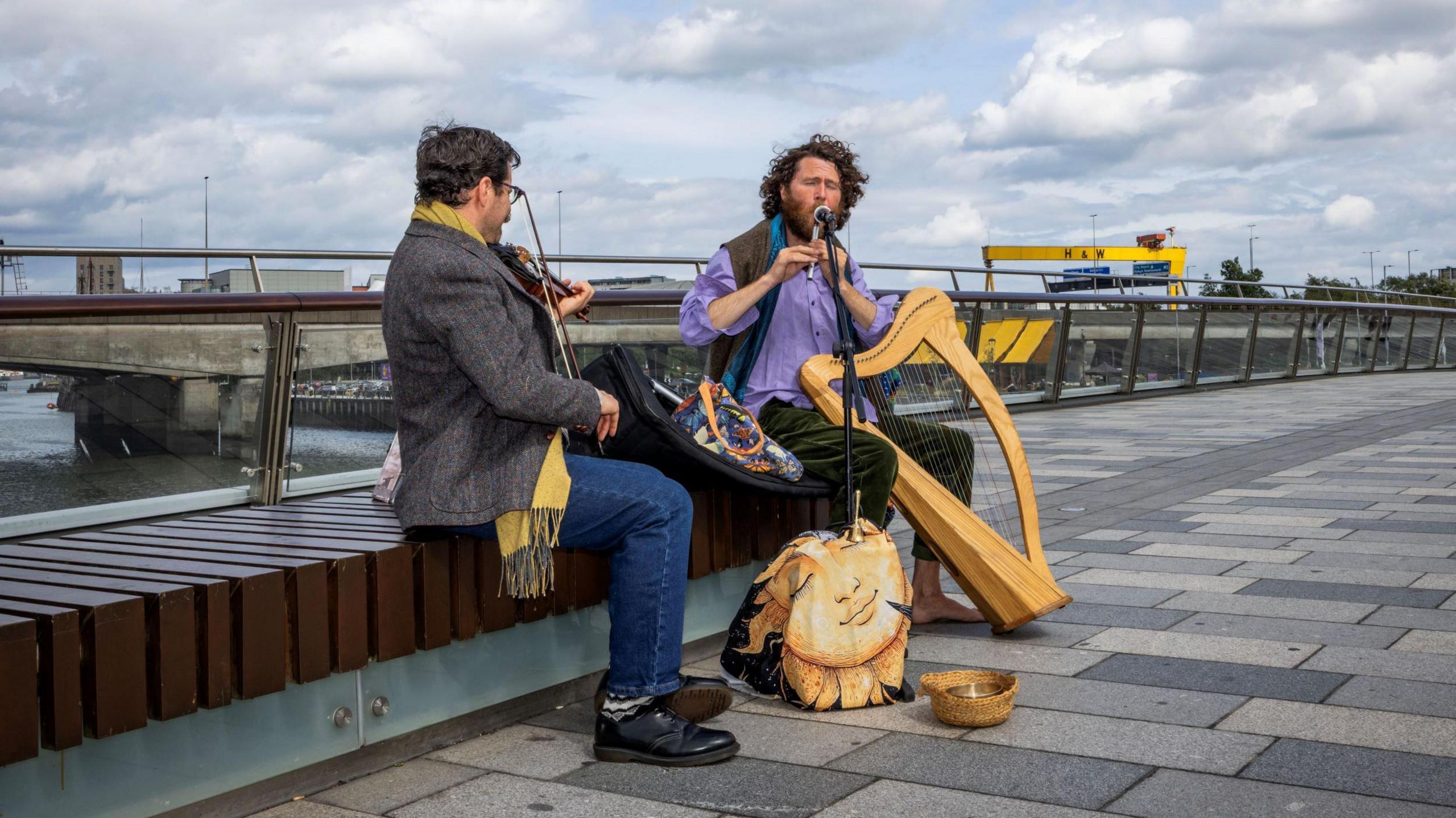 Two men sat near the river in Belfast. One man is playing a tin whistle and the one is playing the fiddle.