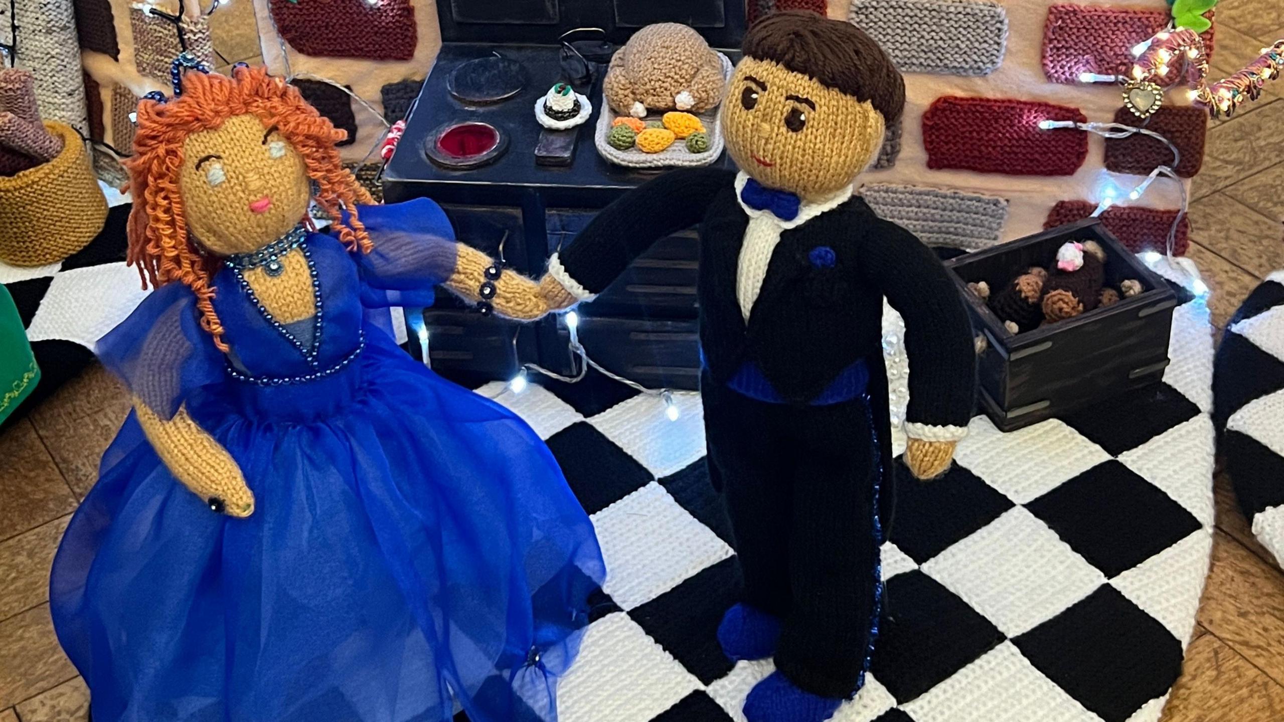 A knitted couple in front of a knitted kitchen, showing food and a turkey. The woman is wearing a blue dress and the man is wearing a tuxedo. They are standing on a black and white floor. 