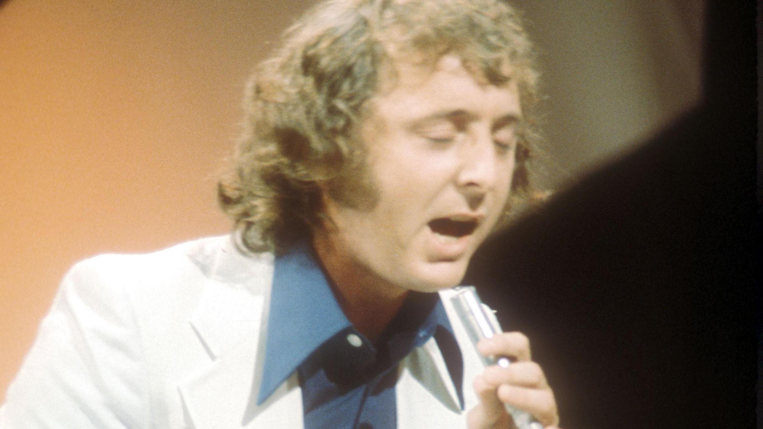 A man with long curly brown hair to his shoulders, sings into a metal tube while wearing a white suit over a blue shirt. 