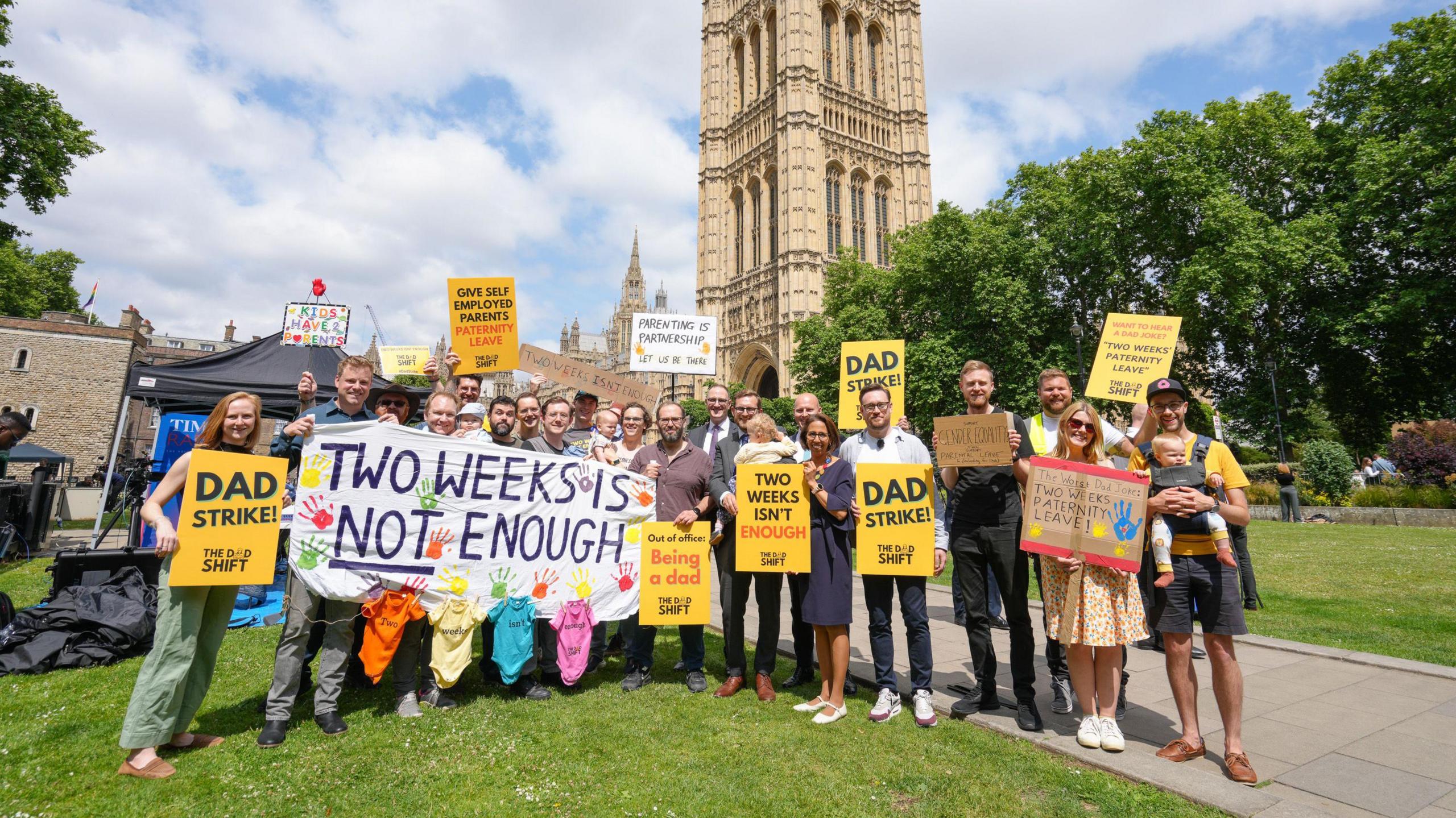 A group of The Dad Shift campaigners holding banners outside the houses of parliament. Some say "dad strike" and some say "two weeks is not enough"