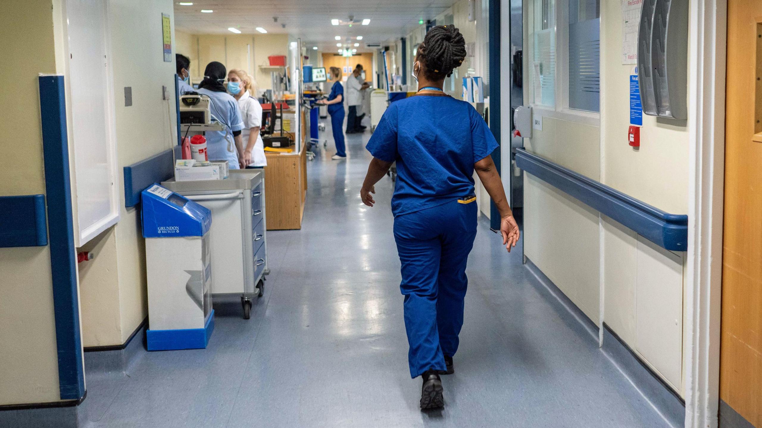 A doctor wearing dark blue scrubs and a face mask, with her back to the camera, walks down a hospital corridor 