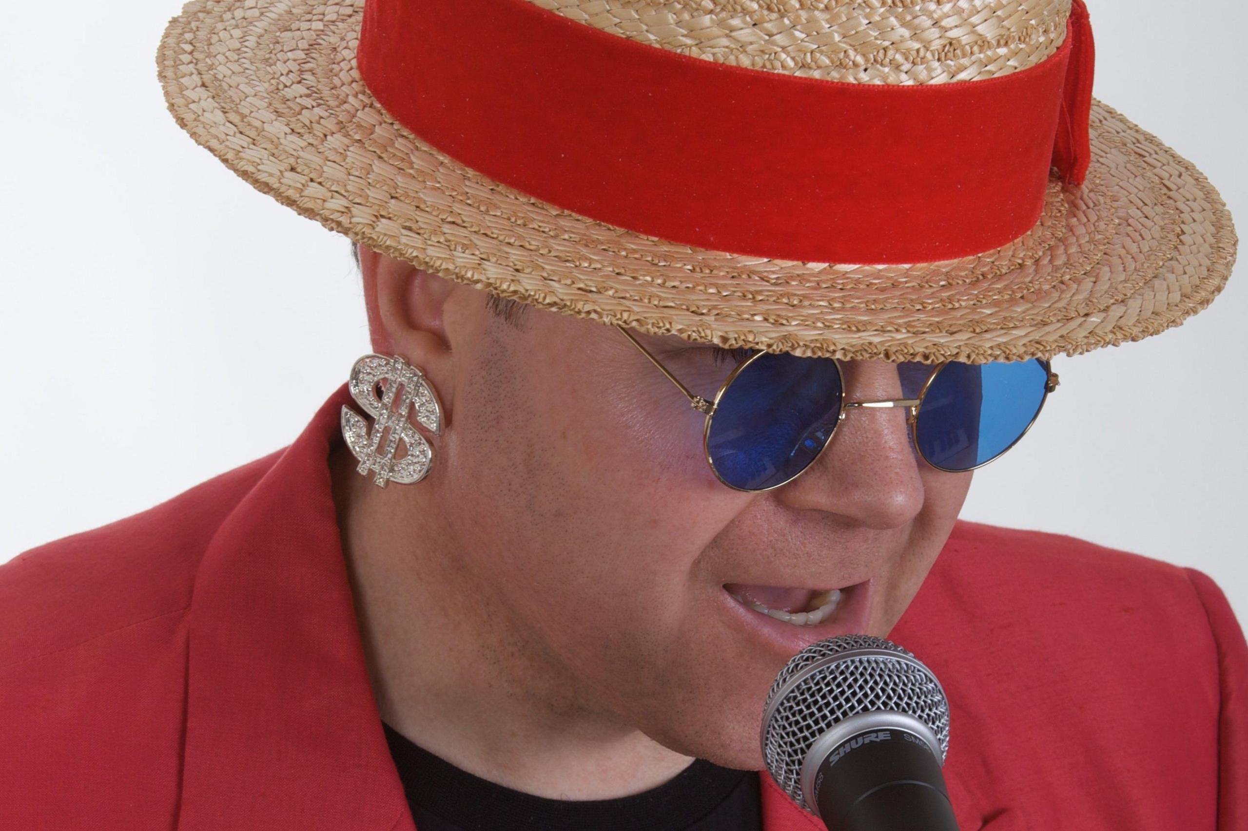 Ultimate Elton, an Elton John tribute act, with a man dressed in a red jacket and hat with a red stripe. He has a dollar sign earring and sunglasses and is singing into a microphone.