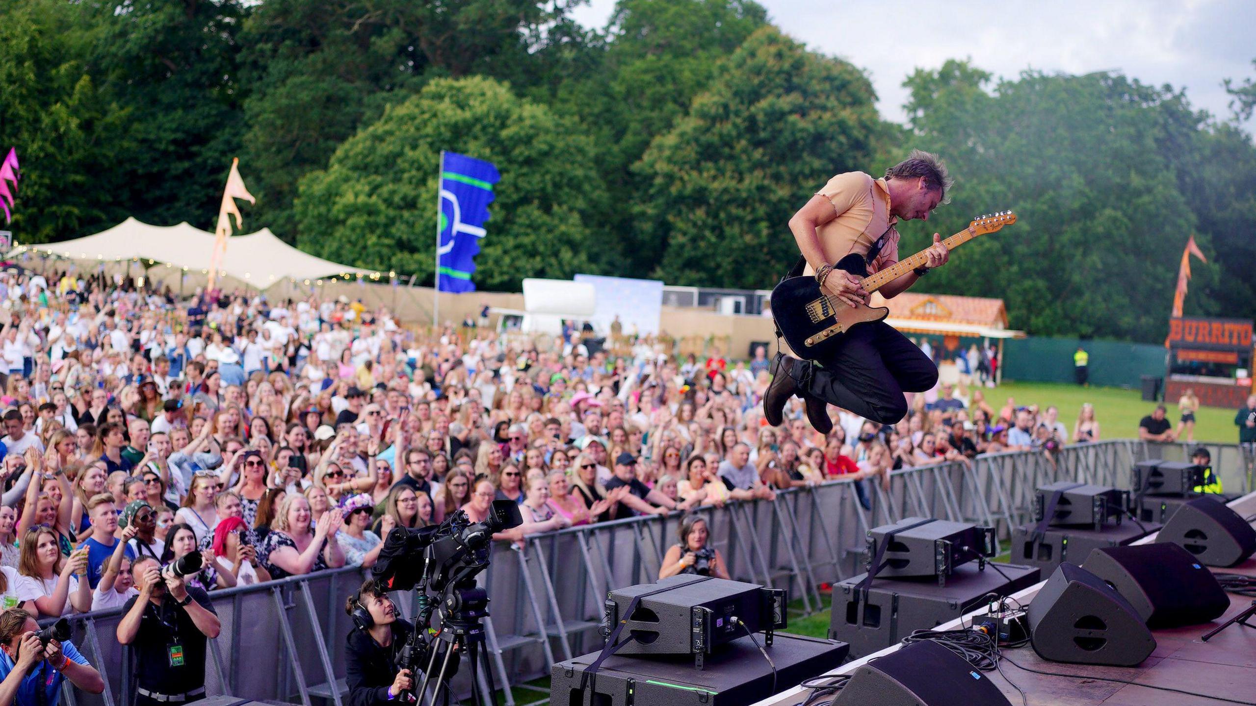 Chesney Hawke in mid air playing a black electric guitar, whilst a crowd looks on in shock in the background