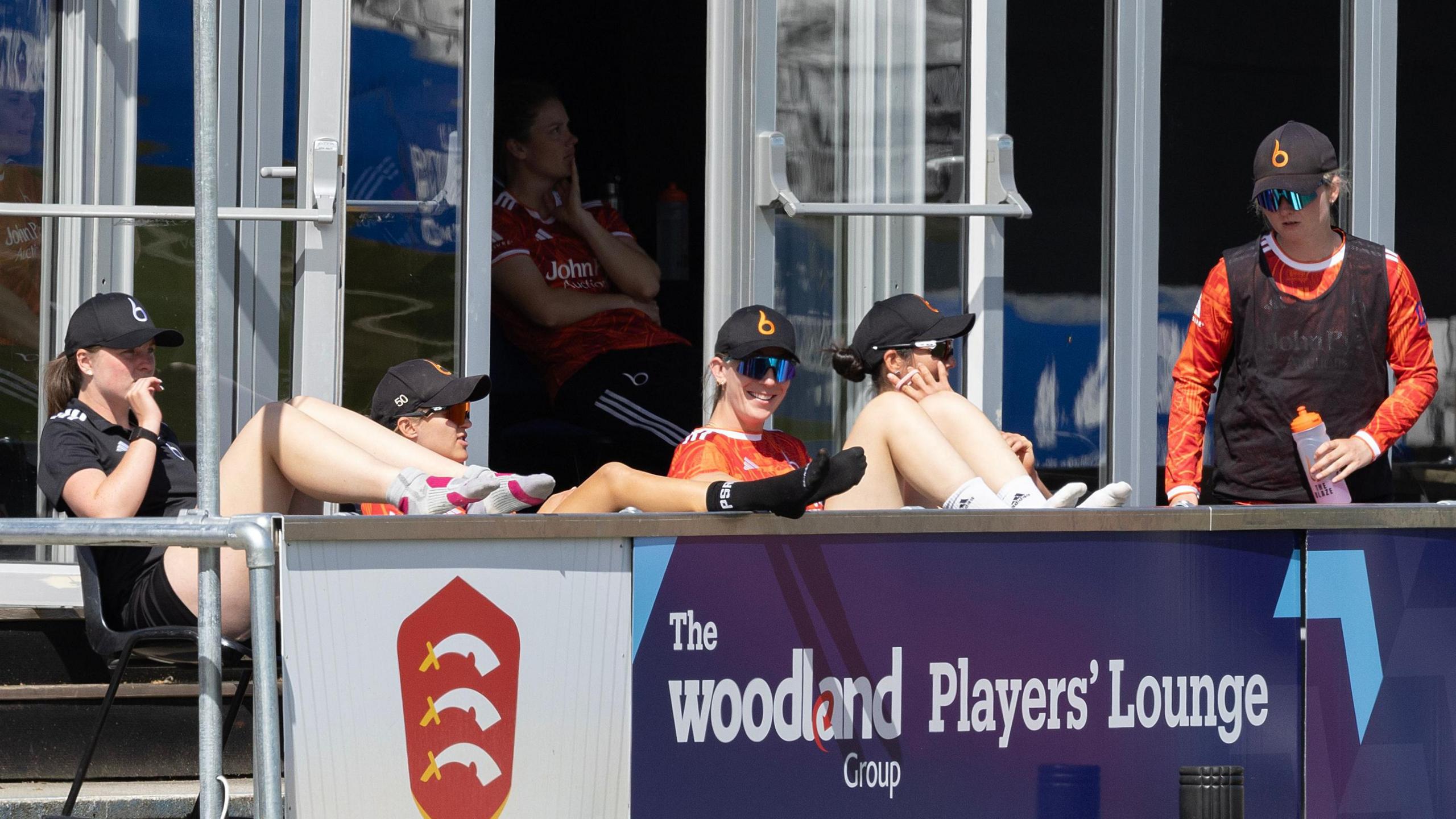 Five Blaze team-mates watch the batting innings at Chelmsford county ground. Four are sitting and observing, whilst one is standing and wearing the 12th player bib.