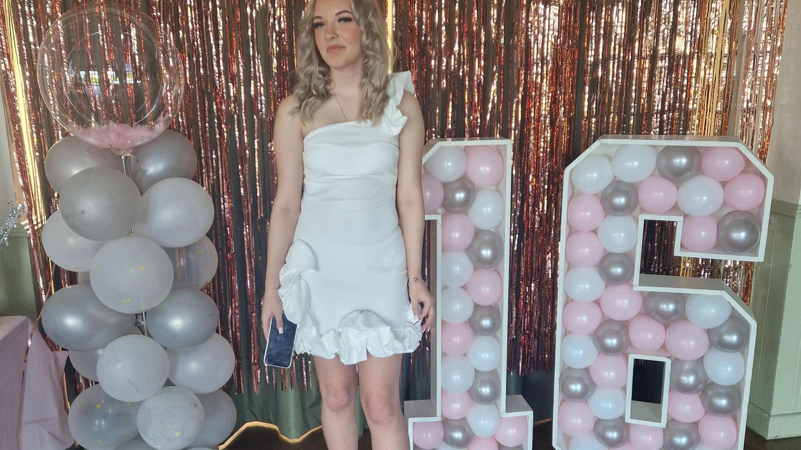 A girl at her 16th birthday party. She is holding a phone and standing next to some balloons. 