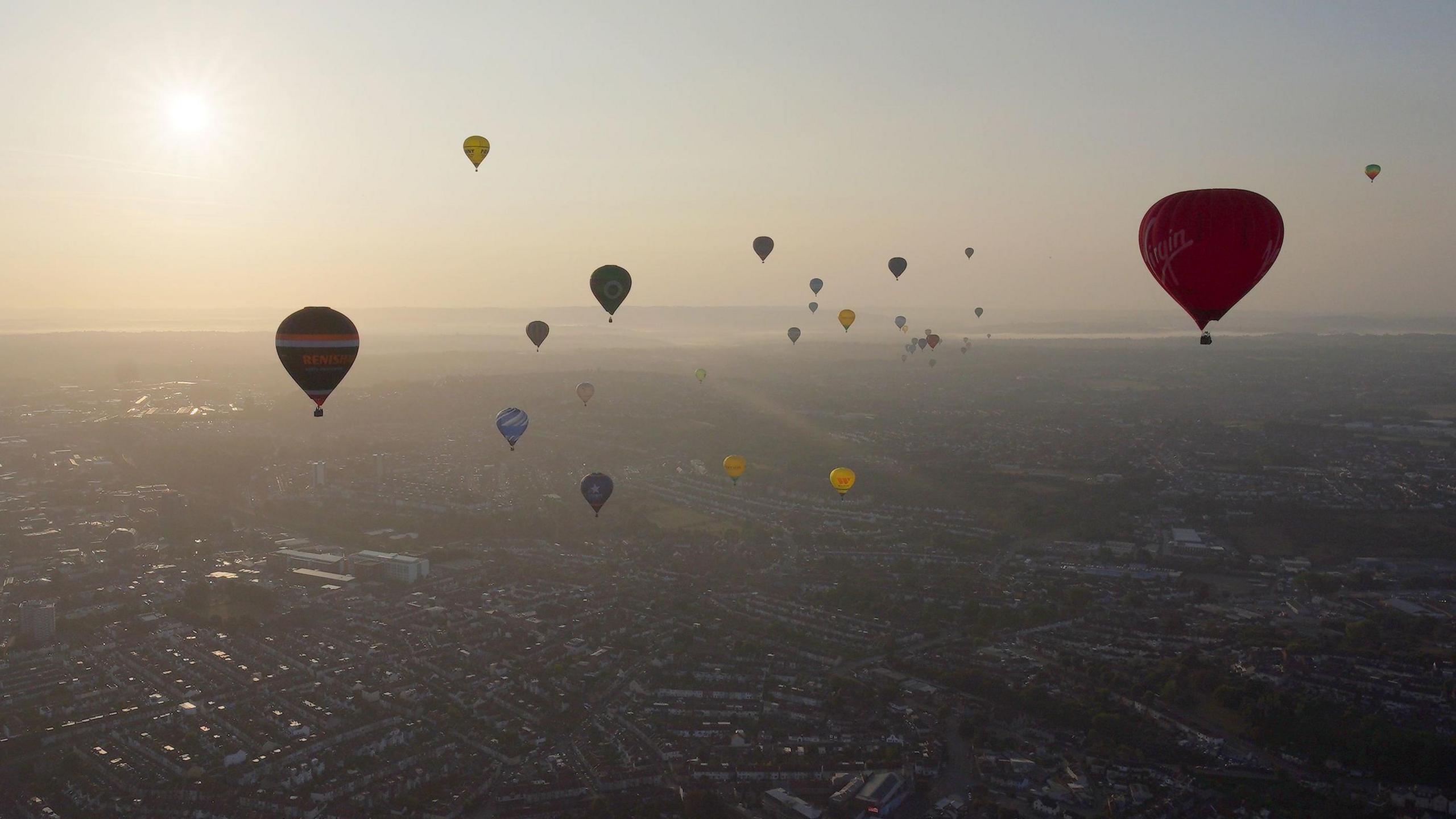 Numerous hot air balloons of all colours fly above Bristol at sunrise. The sky is hazy but clear. Hills and the River Severn can be seen in the distance.