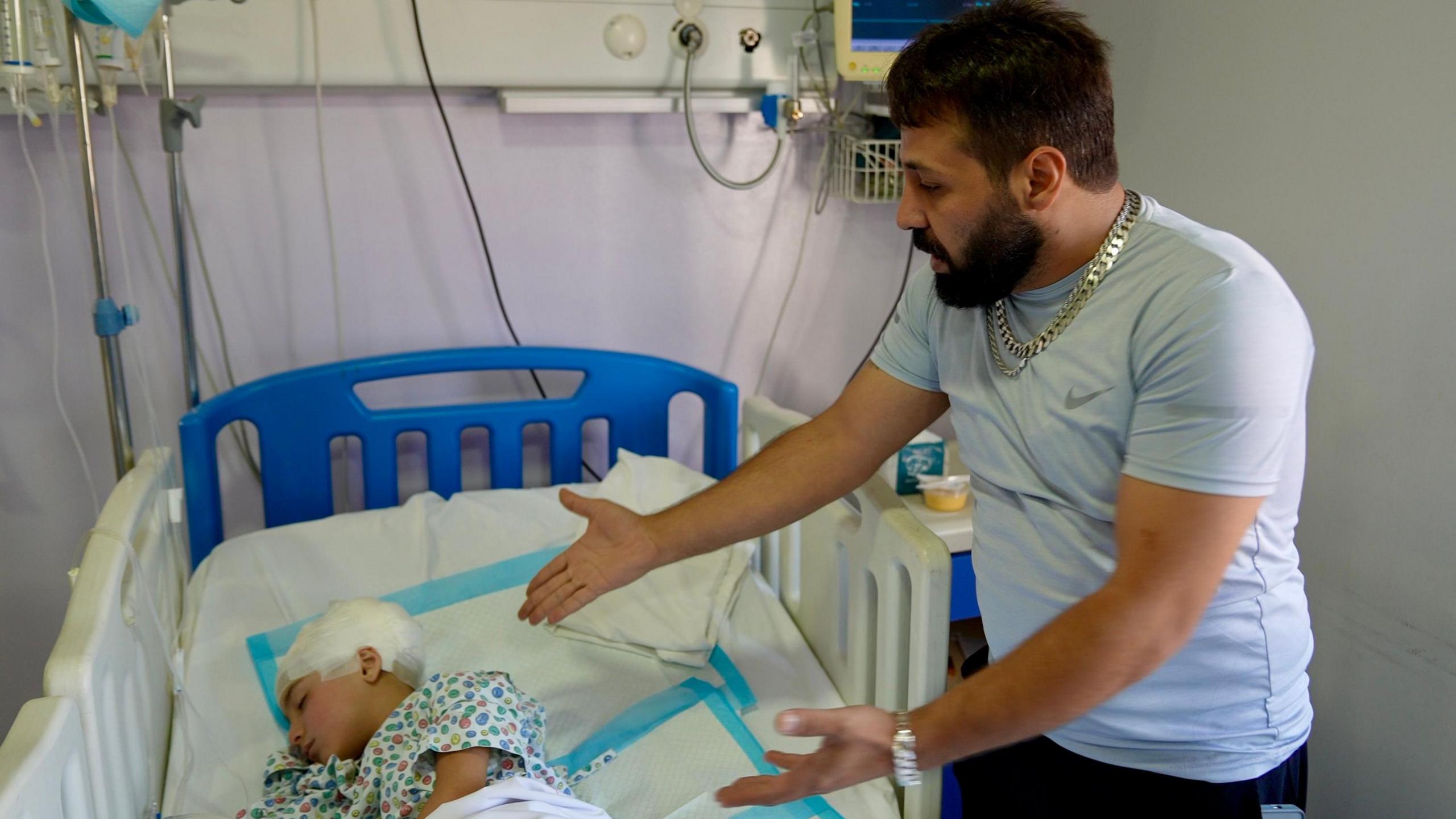 Noor and her father Abdallah in Rayak Hospital, Bekaa Valley, Lebanon - his daughter lies in a bed with her head bandaged and her eyes closed. Standing over her bed, her father wears a chair and gestures to his daughter with with both hands in a gesture of despair. 