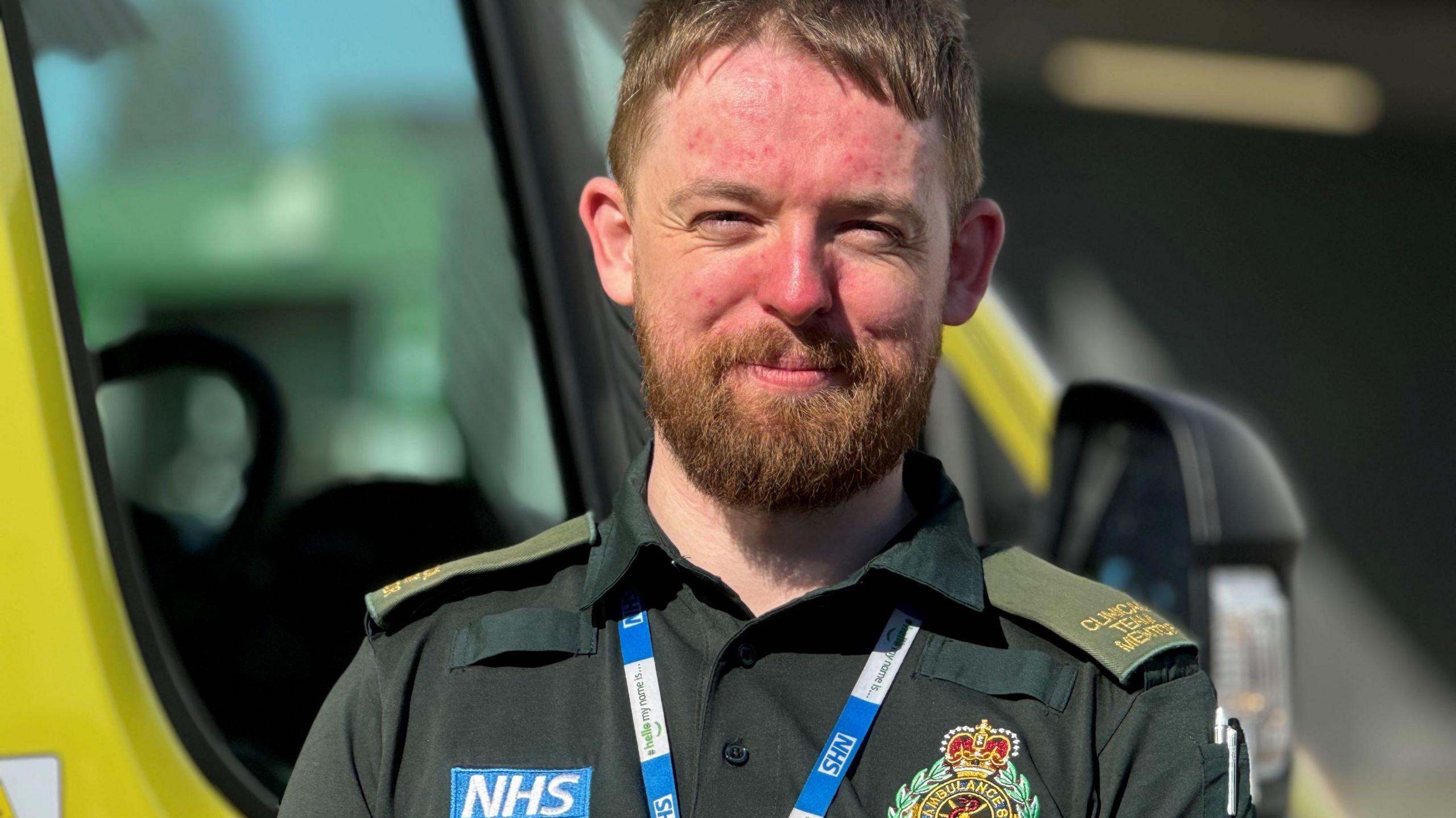 A paramedic wearing a dark green uniform which has an NHS label on. It also has an ambulance badge on it and he is also wearing a blue and white NHS lanyard. The man has auburn hair and and a beard and is smiling at the camera.