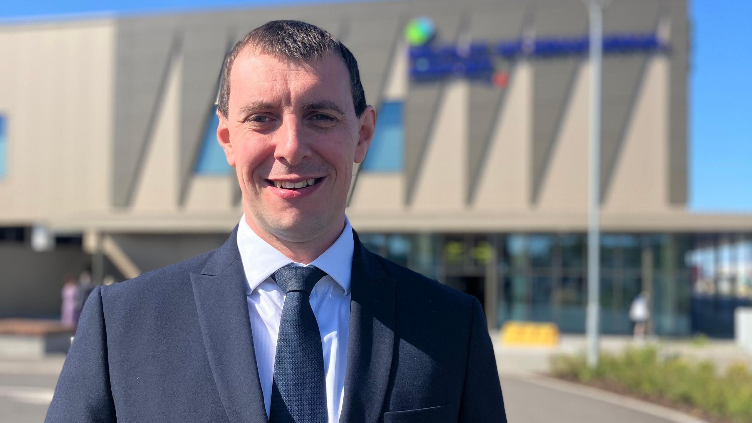 Dan is standing outside the airport in a navy suit with a white shirt underneath. He is wearing a navy tie and has brown short hair. He is smiling at the camera. Behind him is a blurry airport and a blue sky.