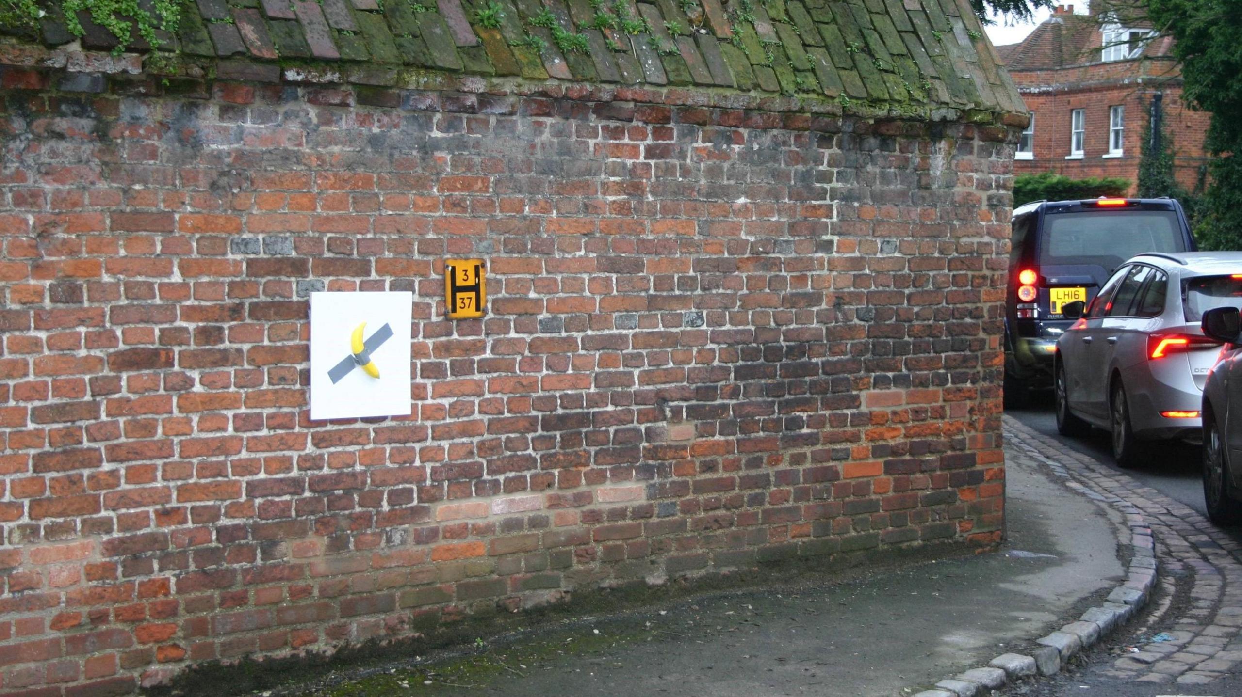 A banana duct-taped to a square white board is affixed to an ageing brick wall on the side of a narrow village street