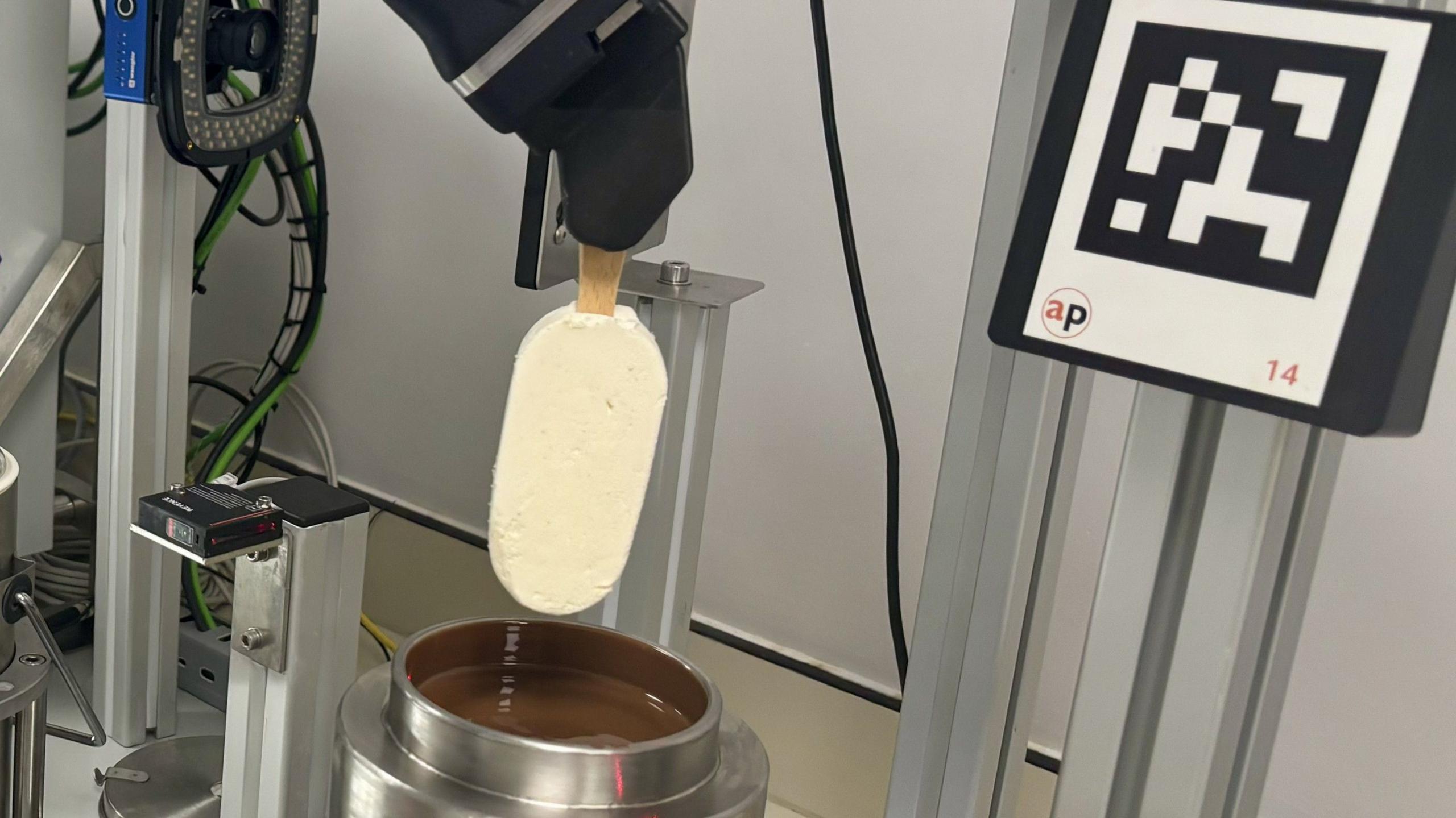 A robot arm holds a Magnum ice cream above a pot of chocolate.