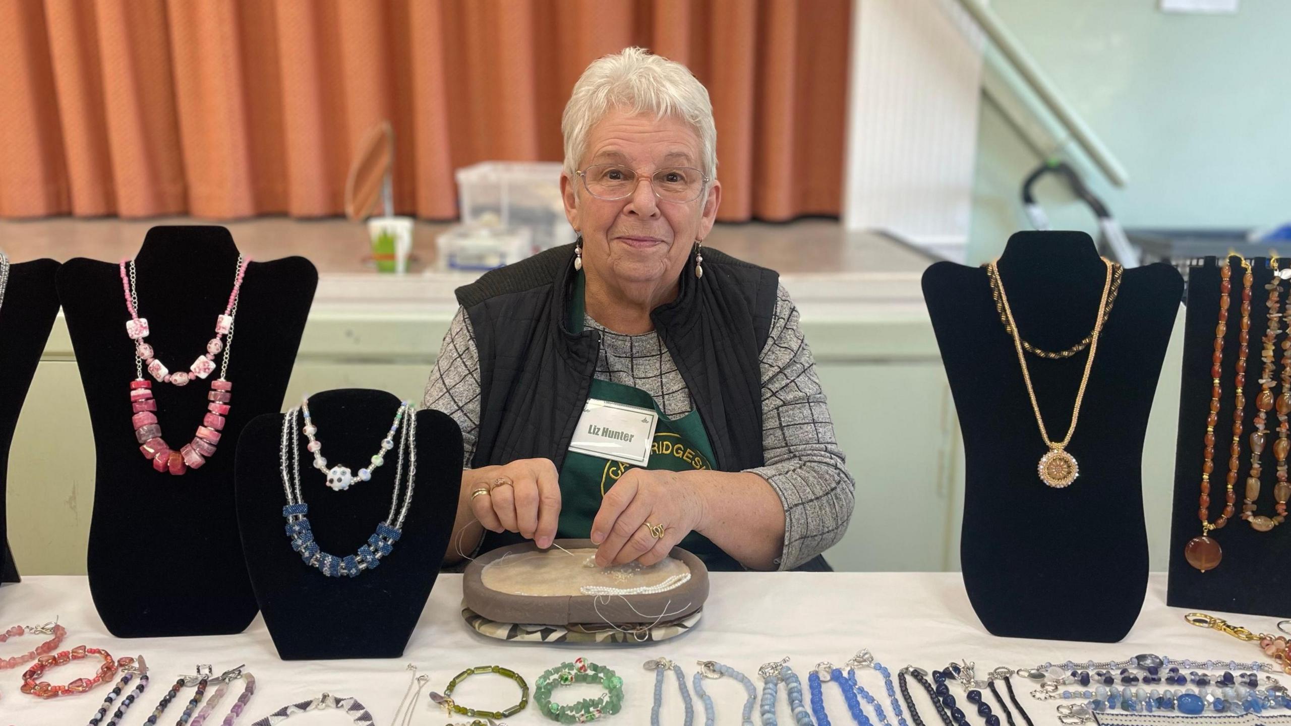 Liz is sitting down and working with a needle and thread, either side of her are busts with necklaces on them, and on the table in front of her is beaded jewellery in many different colours.