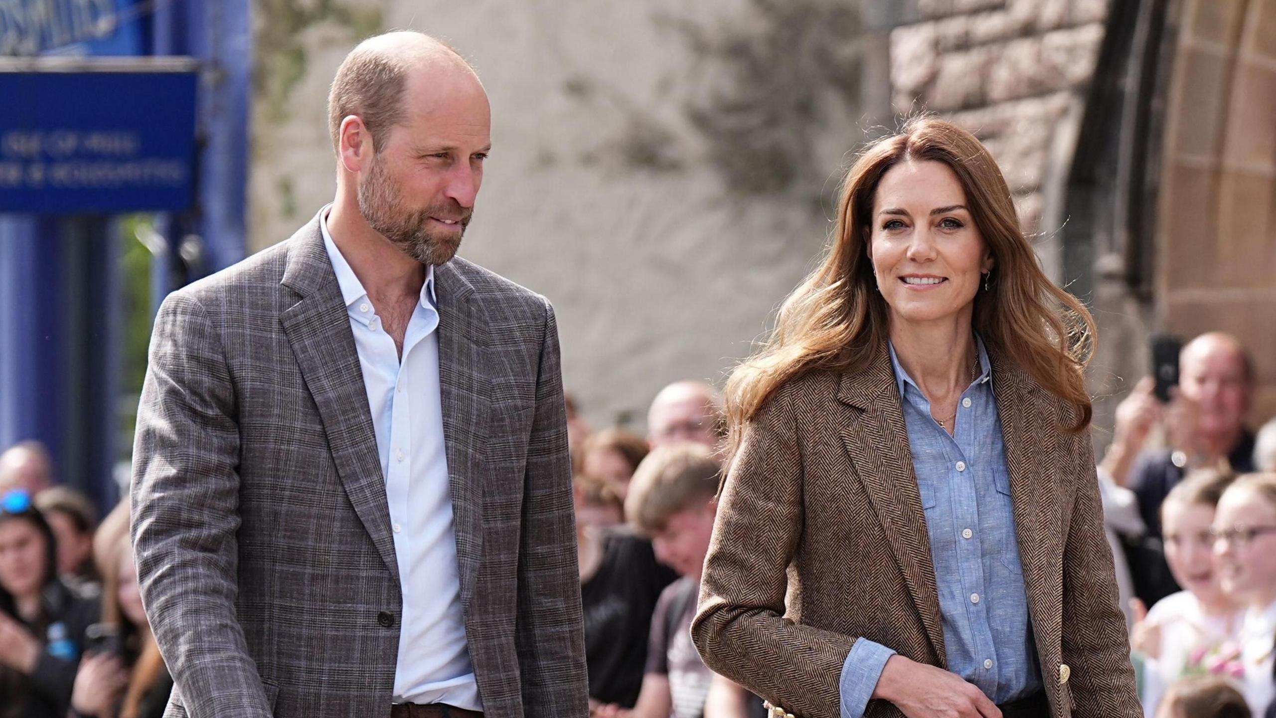 William and Catherine walk with William wearing a grey blazer and blue shirt and Catherine wearing a brown jacket and blue shirt. Catherine is smiling slightly and William has a blank expression.