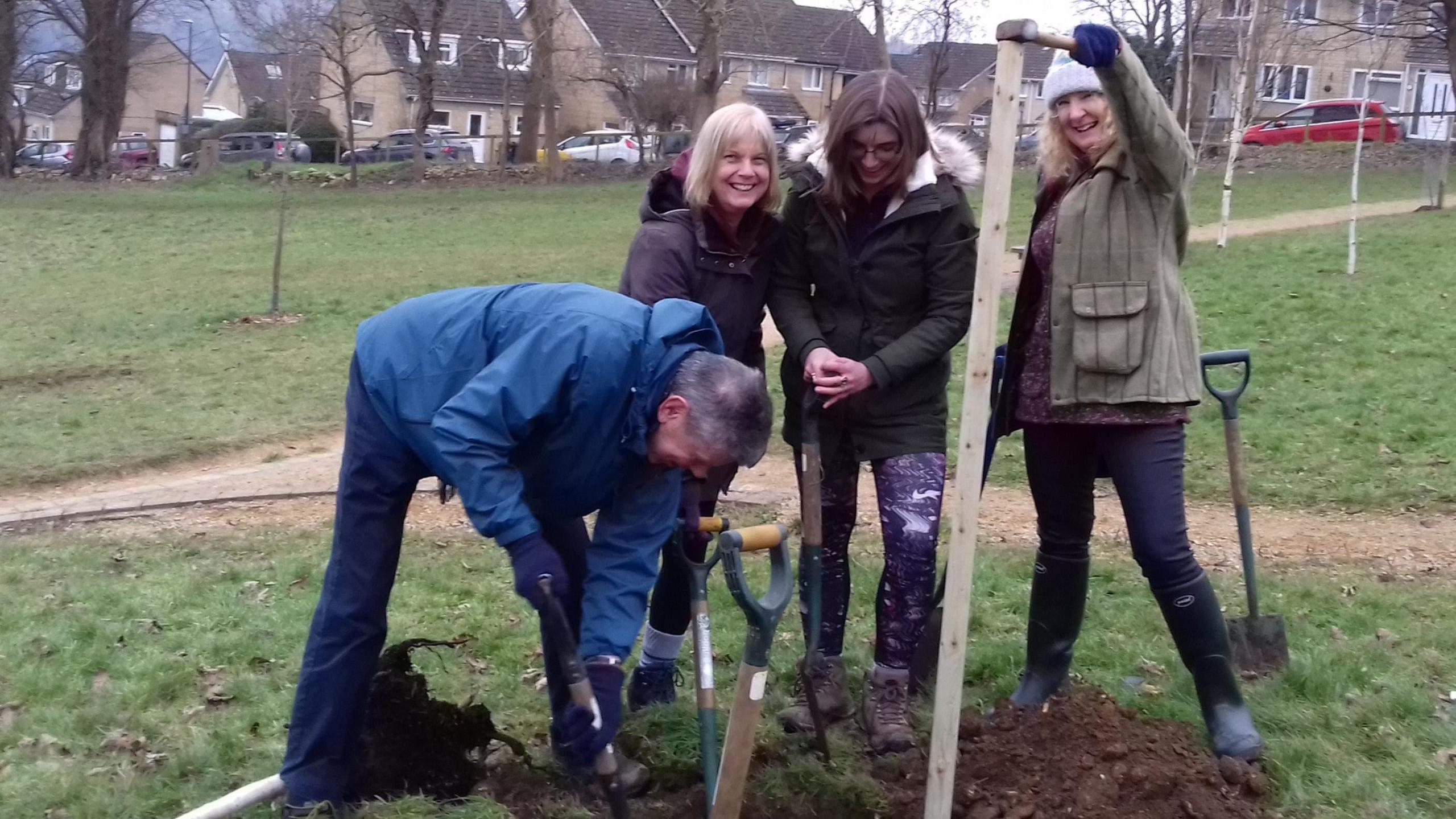 A group of people smiling and using shovels to dig up dirt in the grass with houses in the background in Nailsworth.