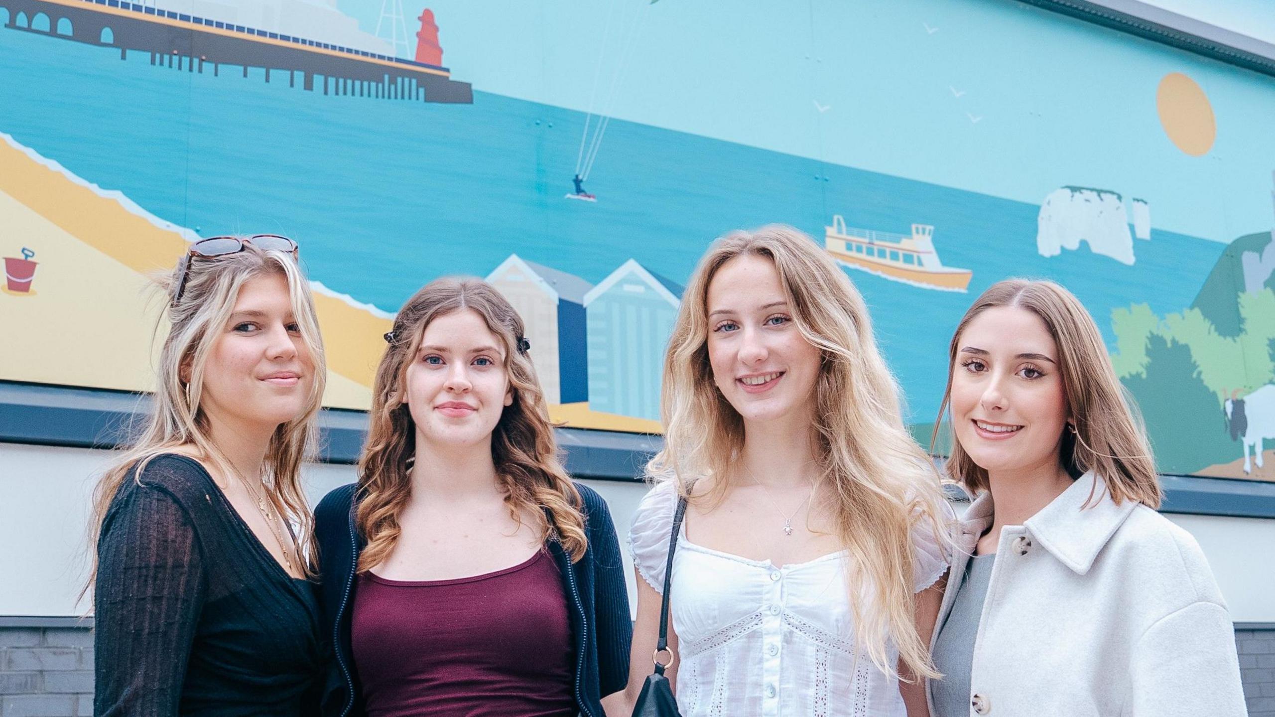Three young women with long fair hair posing in front of a large mural depicting a stylised beach and seaside in block colours with a pier, the white cliffs of Old Harry Rocks, Corfe Castle ruins, a goat, a deckchair and some beach huts.