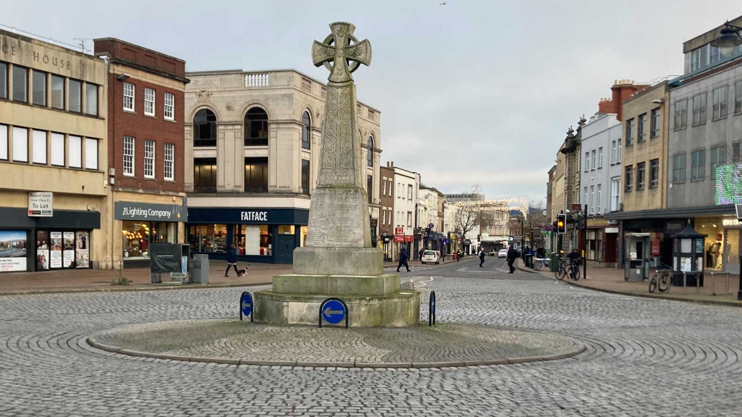 A view of the Market House roundabout on a cobbled street in Taunton town centre with a memorial cross in the middle. In the background are shops and pedestrians crossing the street.